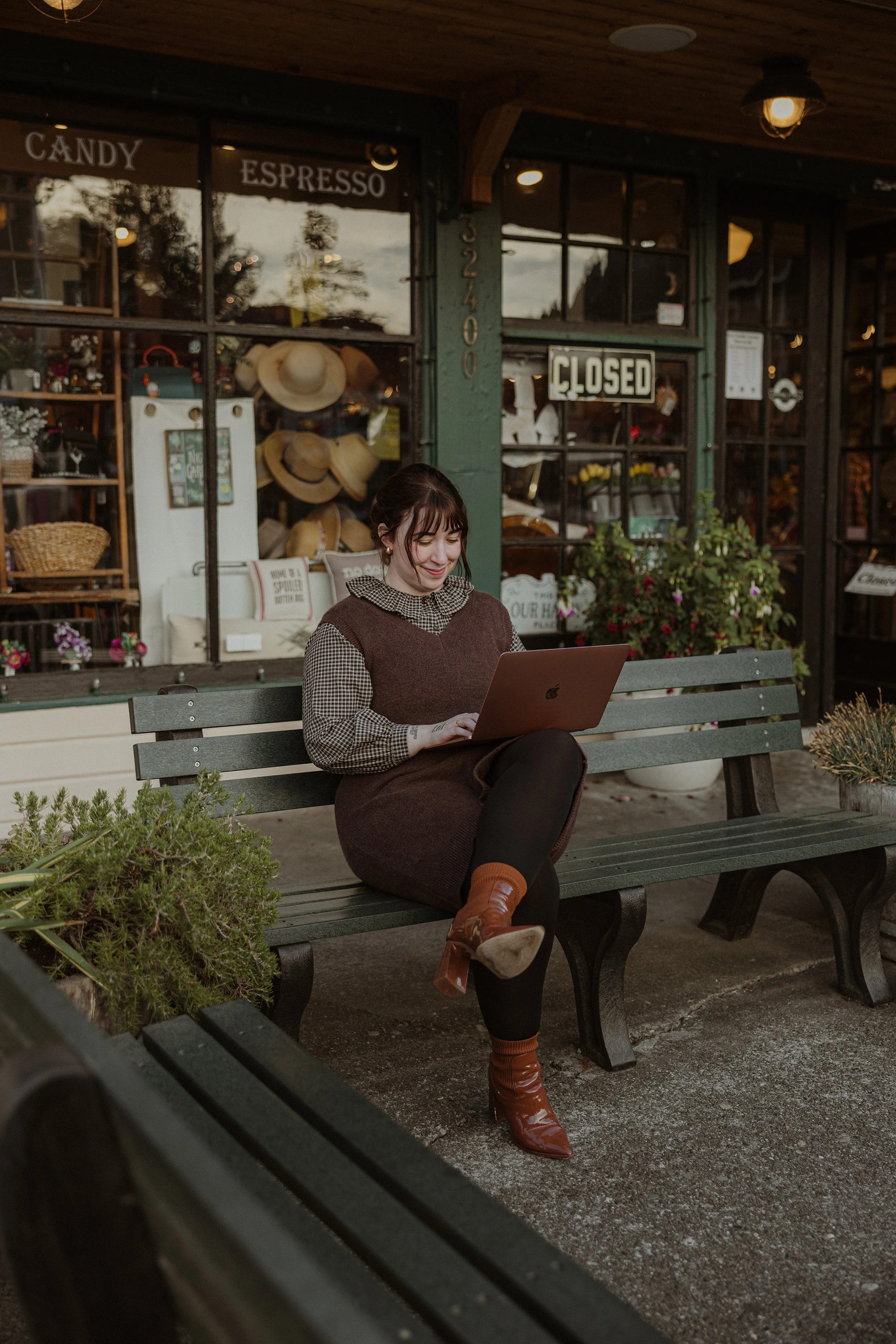 A woman sitting on a park bench using a laptop outside a closed shop with a sign and hats displayed in the window.