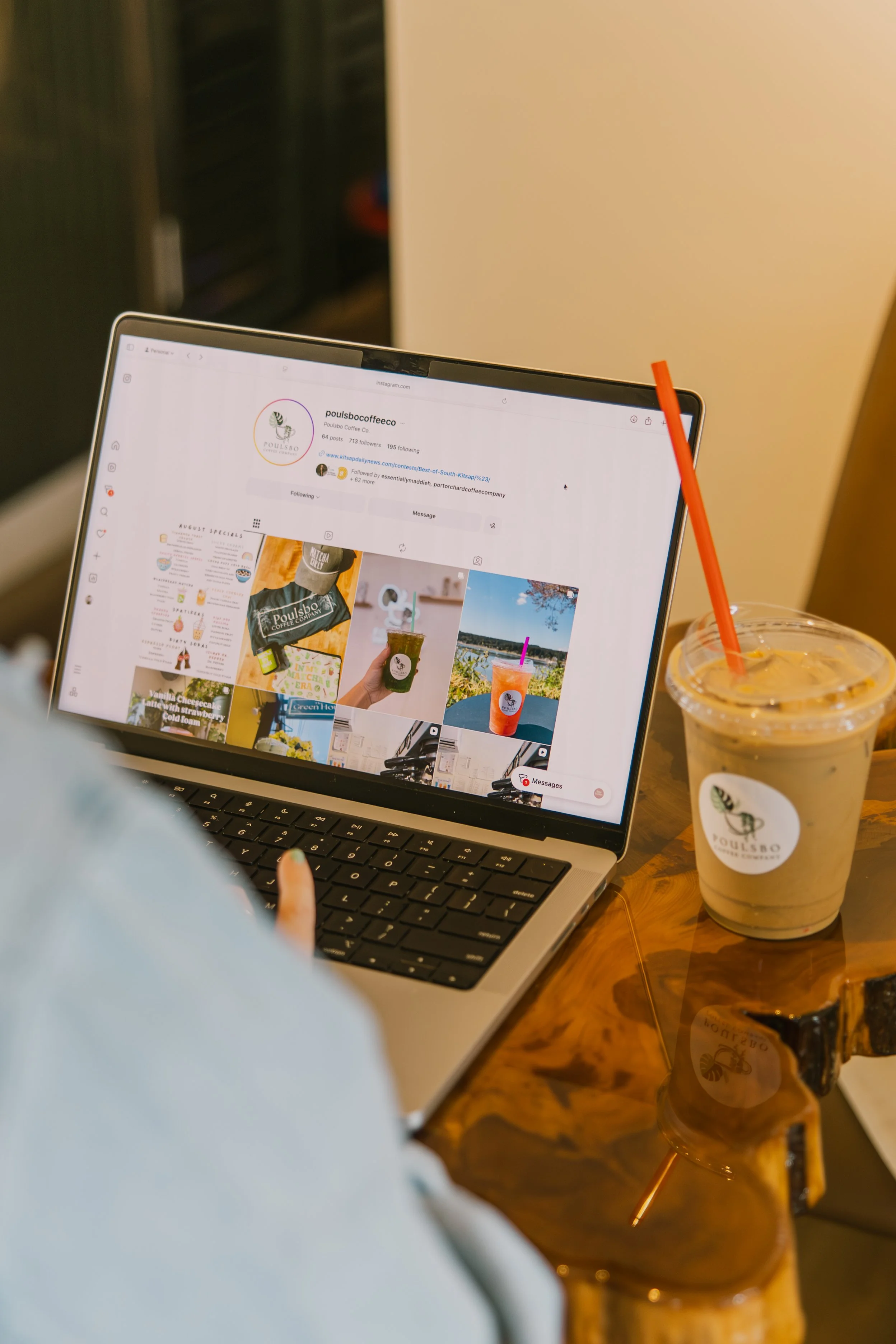 A person using a laptop at a wooden table, with a large iced coffee beverage with a red straw and a branded logo on the cup beside the laptop.
