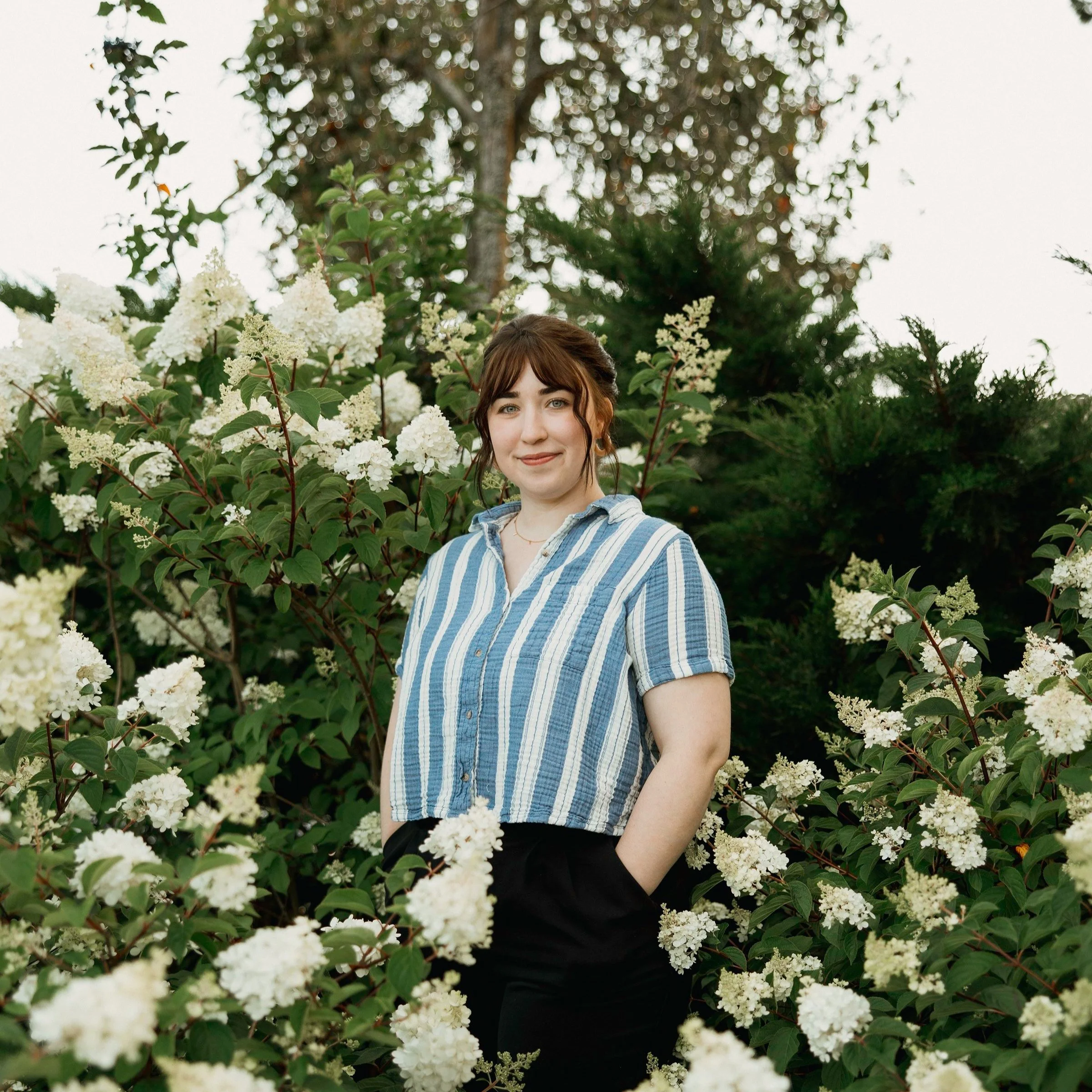 A woman with brown hair and bangs standing outdoors among white flowering bushes and green foliage, smiling at the camera.