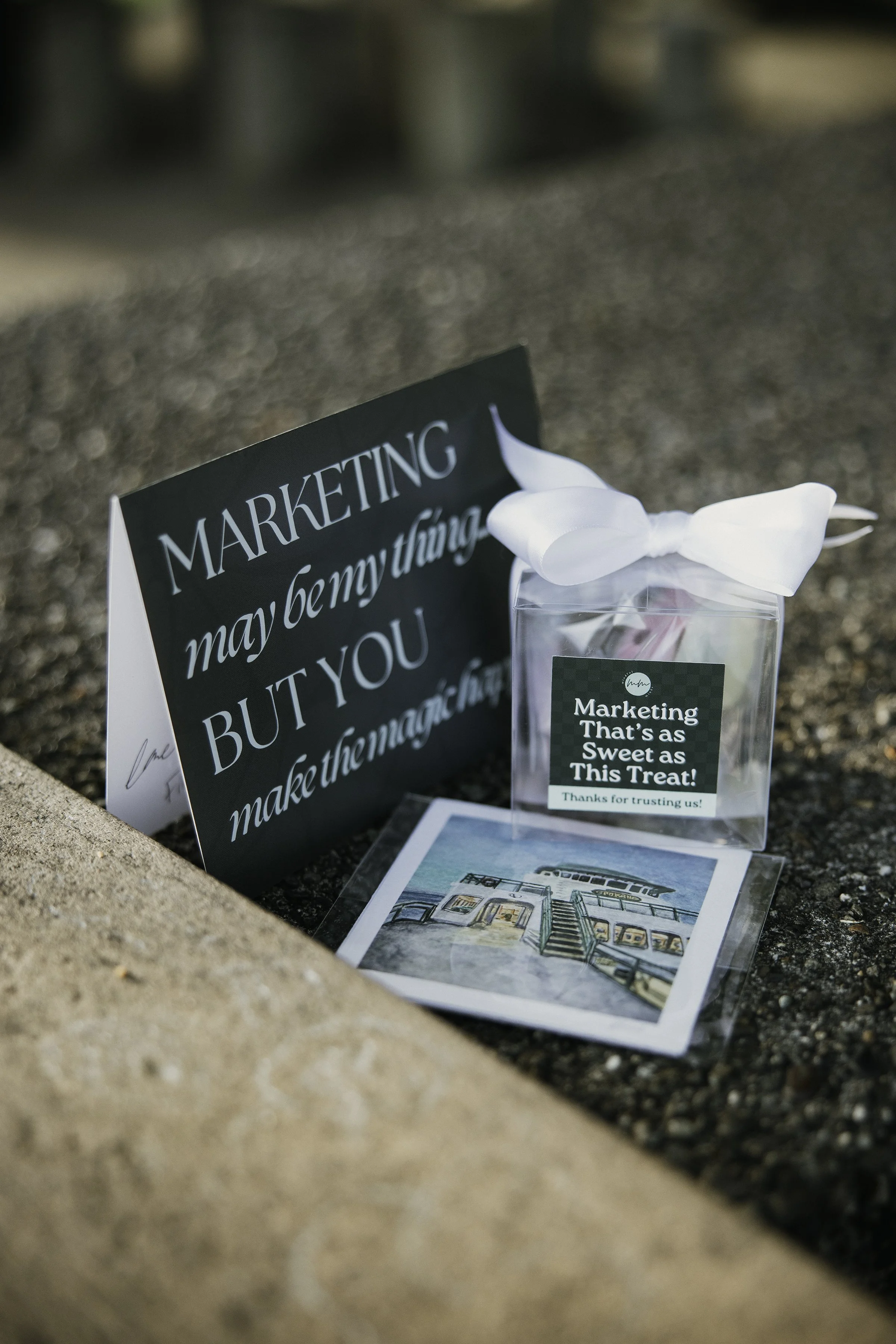 A sign with the quote 'Marketing may be my thing, but you make the magic happen,' a small thank you gift box with a label that reads 'Marketing That's as Sweet as This Treat!', and a postcard with an illustration of a lighthouse and a building, all placed on the ground.