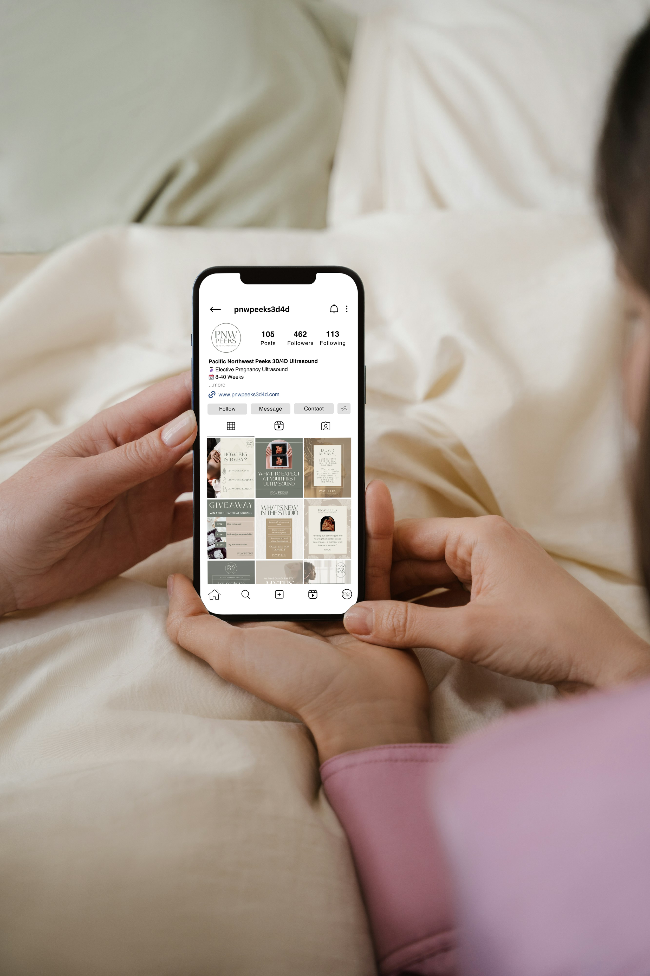Person holding a smartphone displaying an Instagram profile for Pacific Northwest Peaks 3D/4D Ultrasound, with a bed and beige blanket in the background.