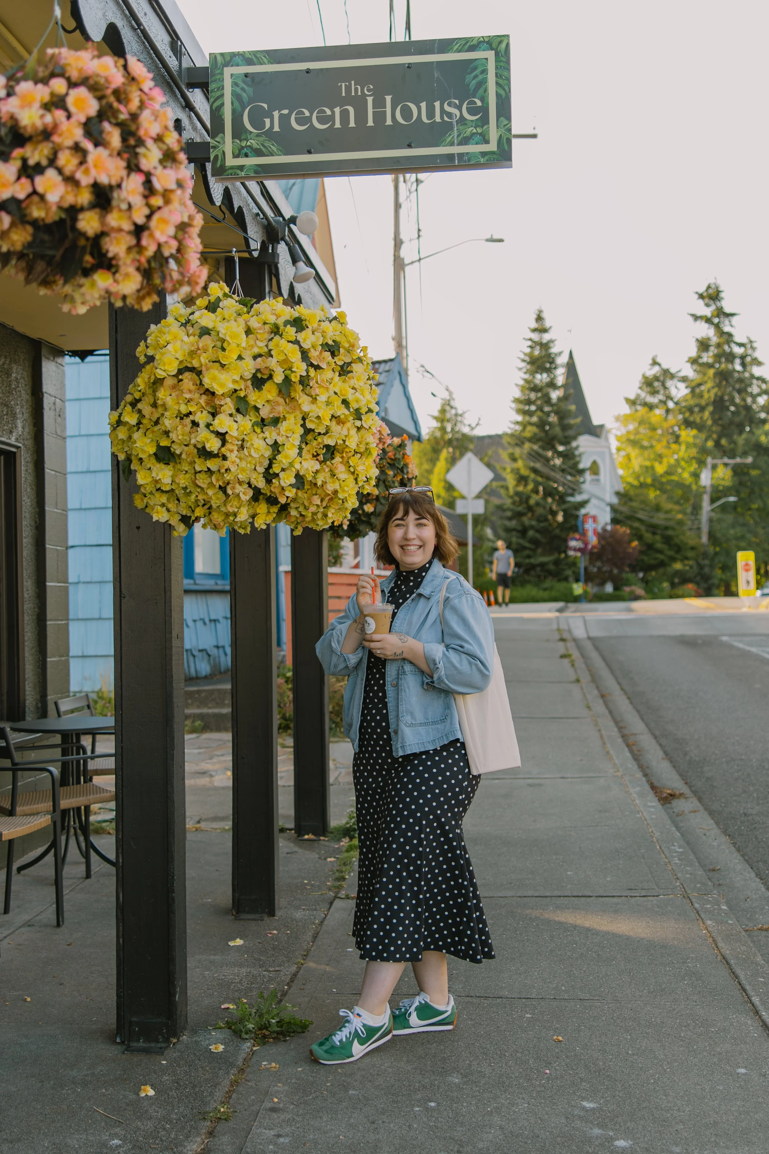 A woman in a black polka dot dress, denim jacket, and green sneakers holding iced coffee, standing on a sidewalk in front of a shop with hanging flower baskets and a sign that says 'The Green House.'