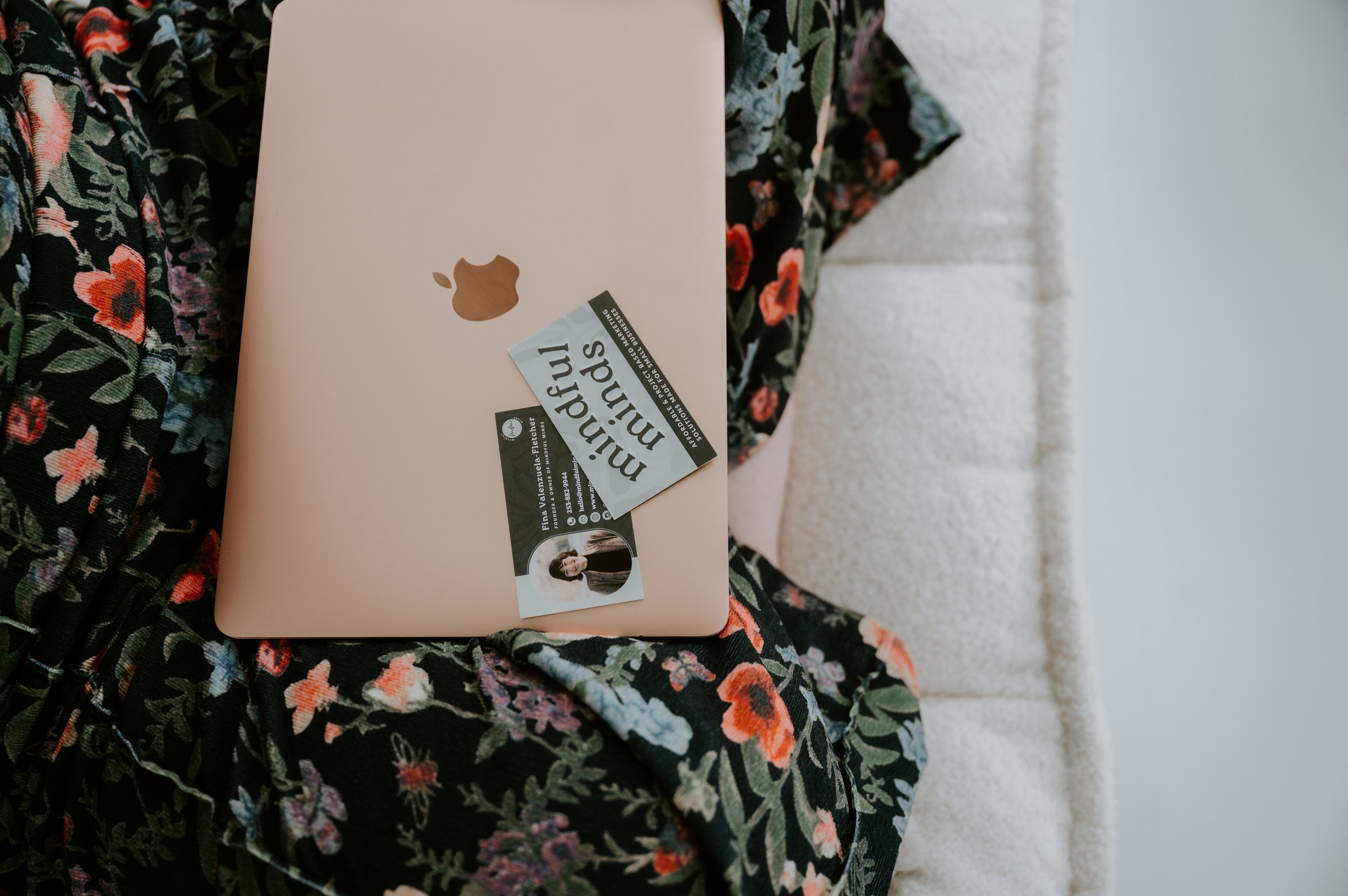 A closed rose gold Apple MacBook laptop with two business cards resting on top of it, placed on a person wearing a black dress with a floral pattern of red, pink, orange, and white flowers. The background includes a cream-colored textured wall.