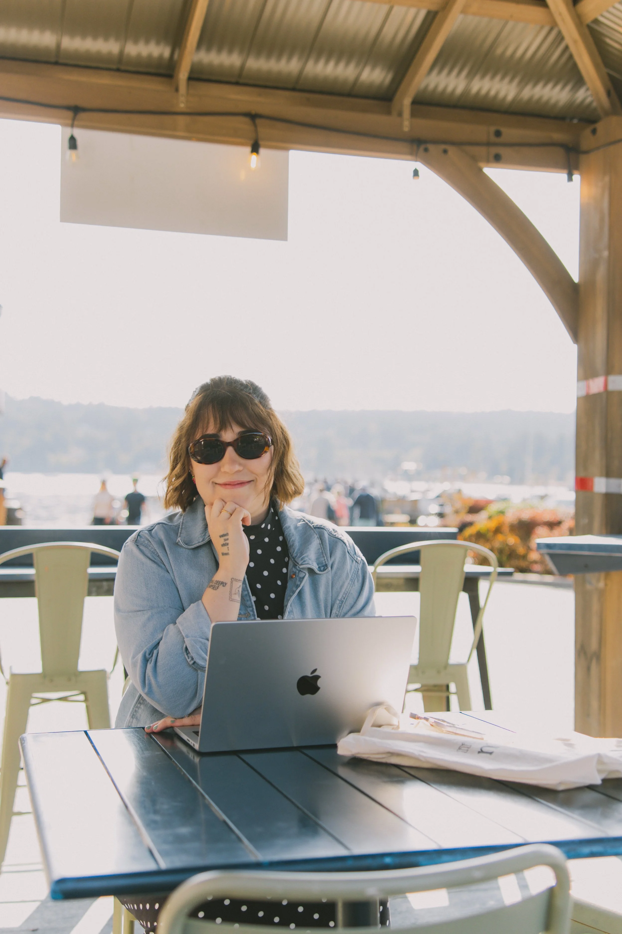A woman with sunglasses sitting at a table with a laptop, looking at the camera, outdoors near a body of water, with a wooden pavilion overhead.