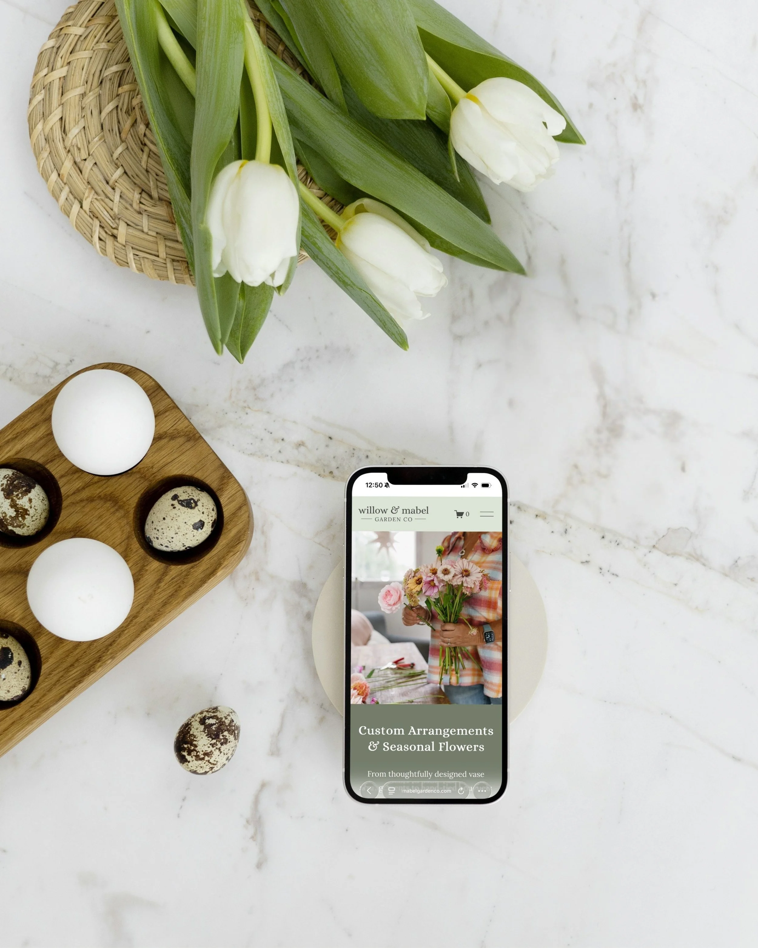 A smartphone displaying a floral arrangement website on a marble surface with white tulips, eggs, and quail eggs nearby.