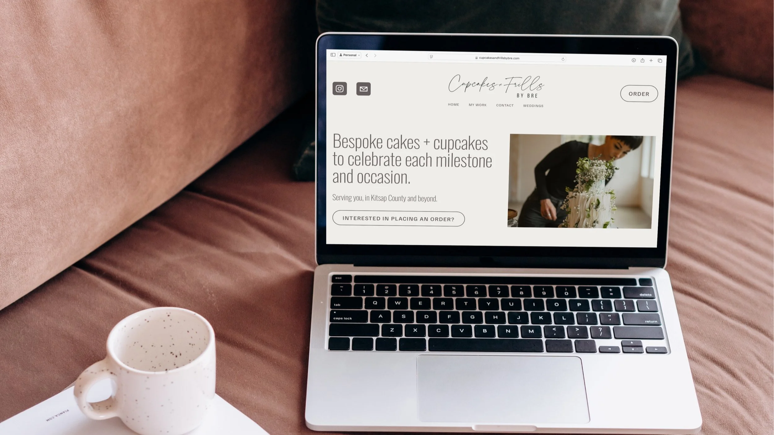 A silver laptop displaying a website for a cake business, placed on a pinkish sofa. Next to the laptop, there is a white speckled ceramic mug and a white notebook or planner.