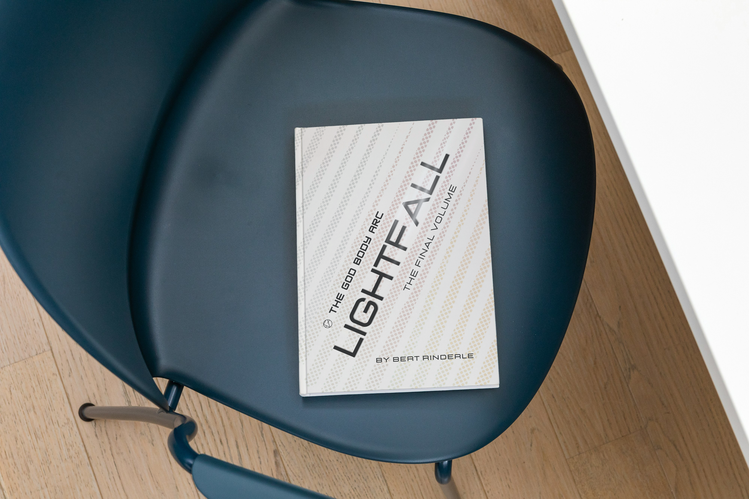 A dark blue chair with a book titled "The God Body Are God Shall: The Final Volume" by Bert Rinderle resting on it. The chair is next to a white table, and the floor is made of light-colored wood.