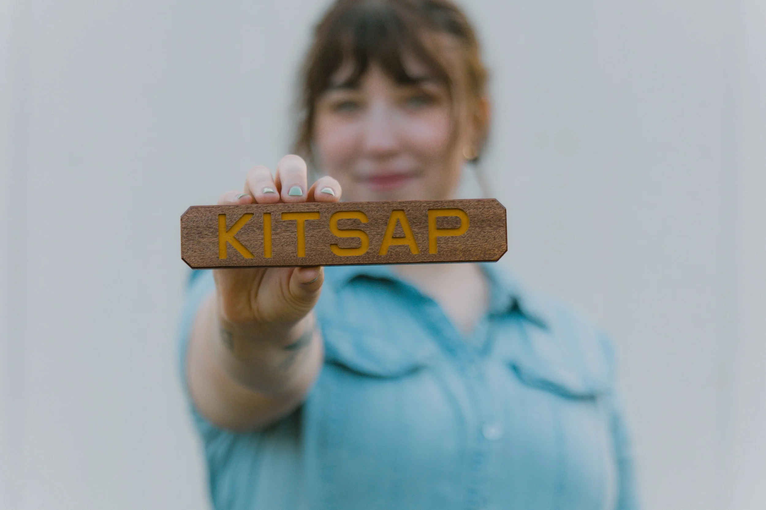 A woman with short brown hair and light blue top holding a wooden sign with yellow letters that spell "KITSAP" in front of her face, against a plain light background.