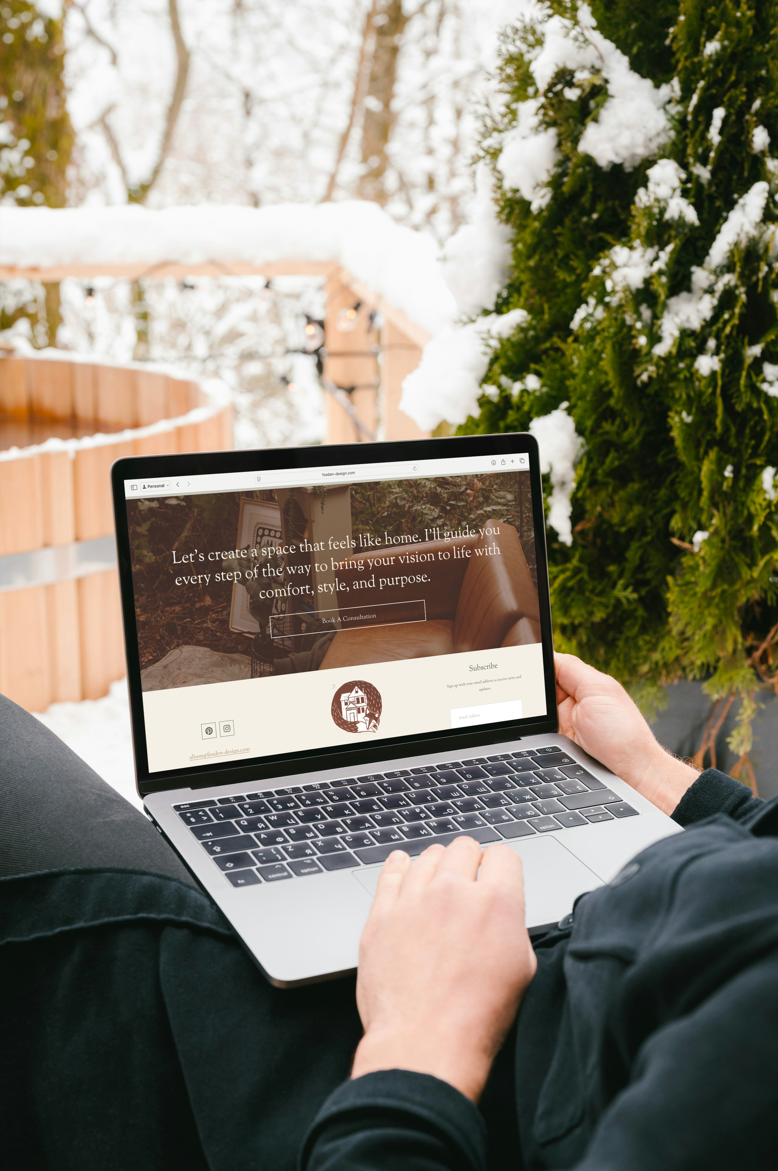 Person using a laptop outdoors on a snowy patio with green trees and a wooden fence in the background.