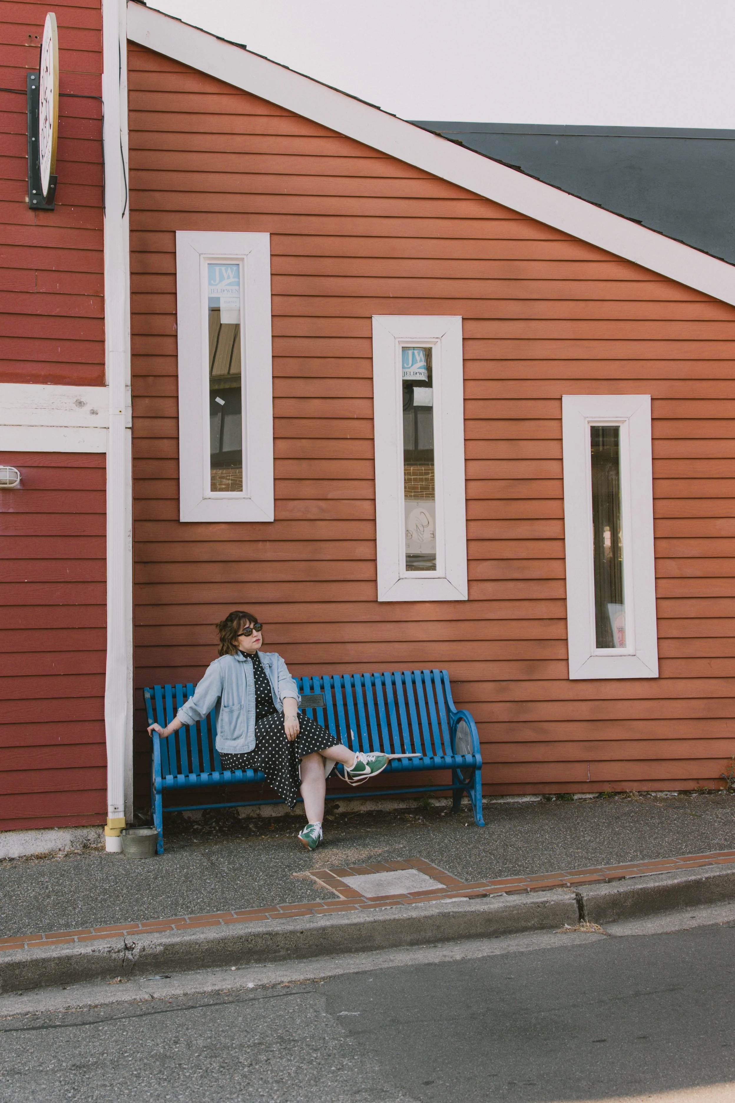 A woman with sunglasses, a Polka dot dress, and a light denim jacket sitting on a blue park bench in front of a red wooden building with white-framed windows and a diagonal roof.