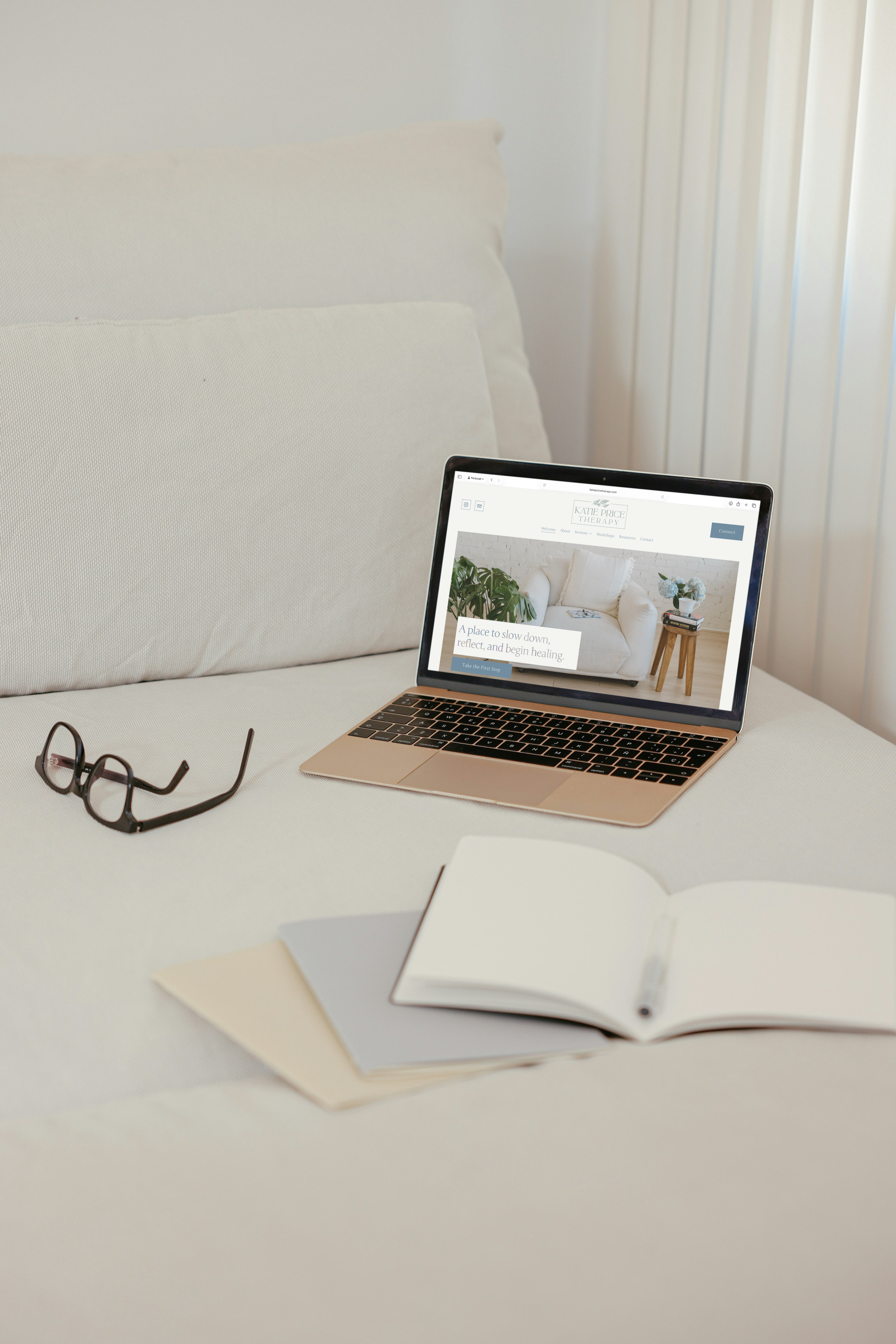 A laptop on a bed displaying a website for Katie Price Therapy, with a pair of glasses, an open notebook, and closed folders nearby.
