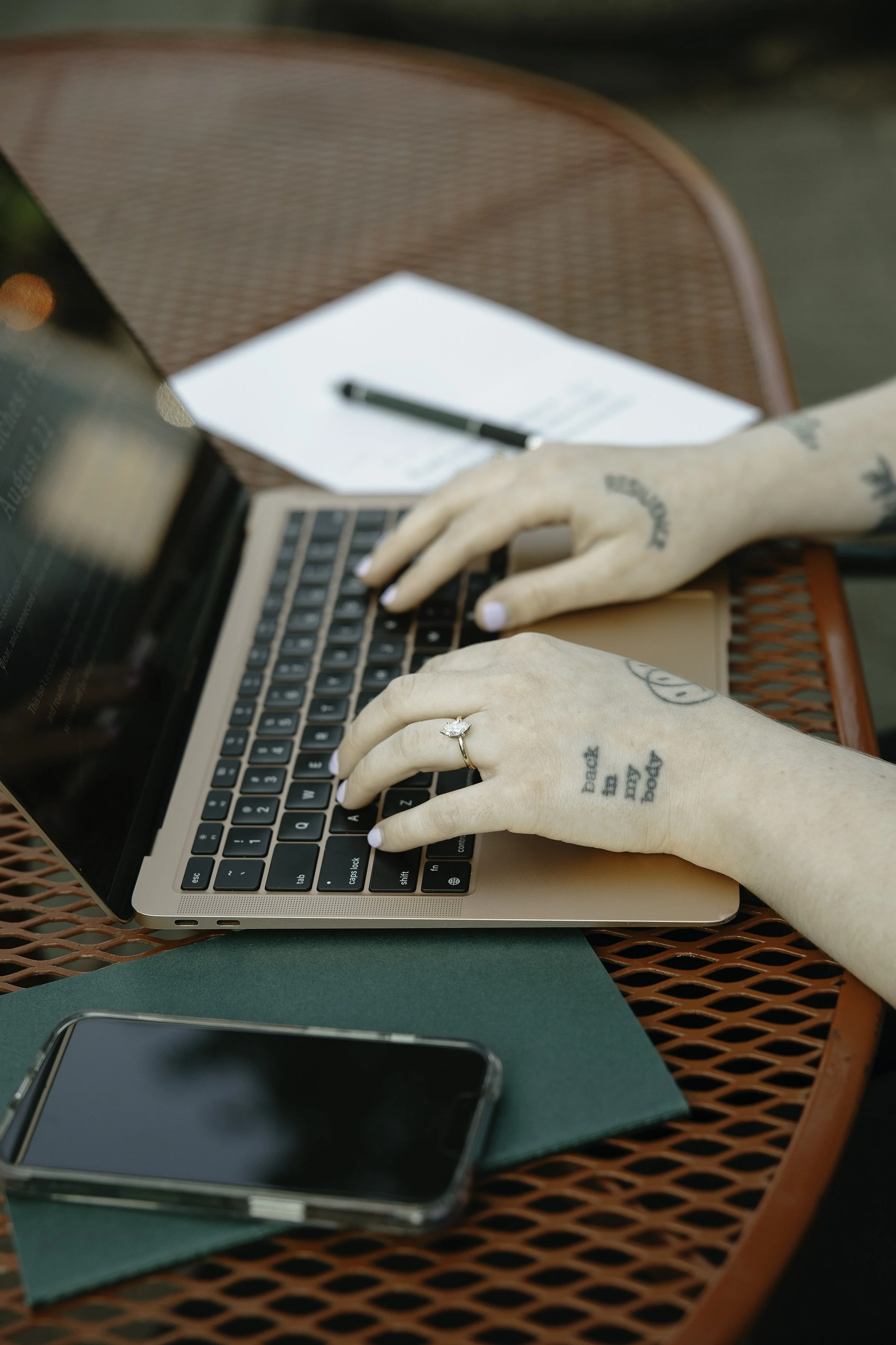 A person's hands typing on a laptop keyboard, with a ring on the left hand's ring finger, tattoos on the hands, and a green notebook and a smartphone on a red metal table.