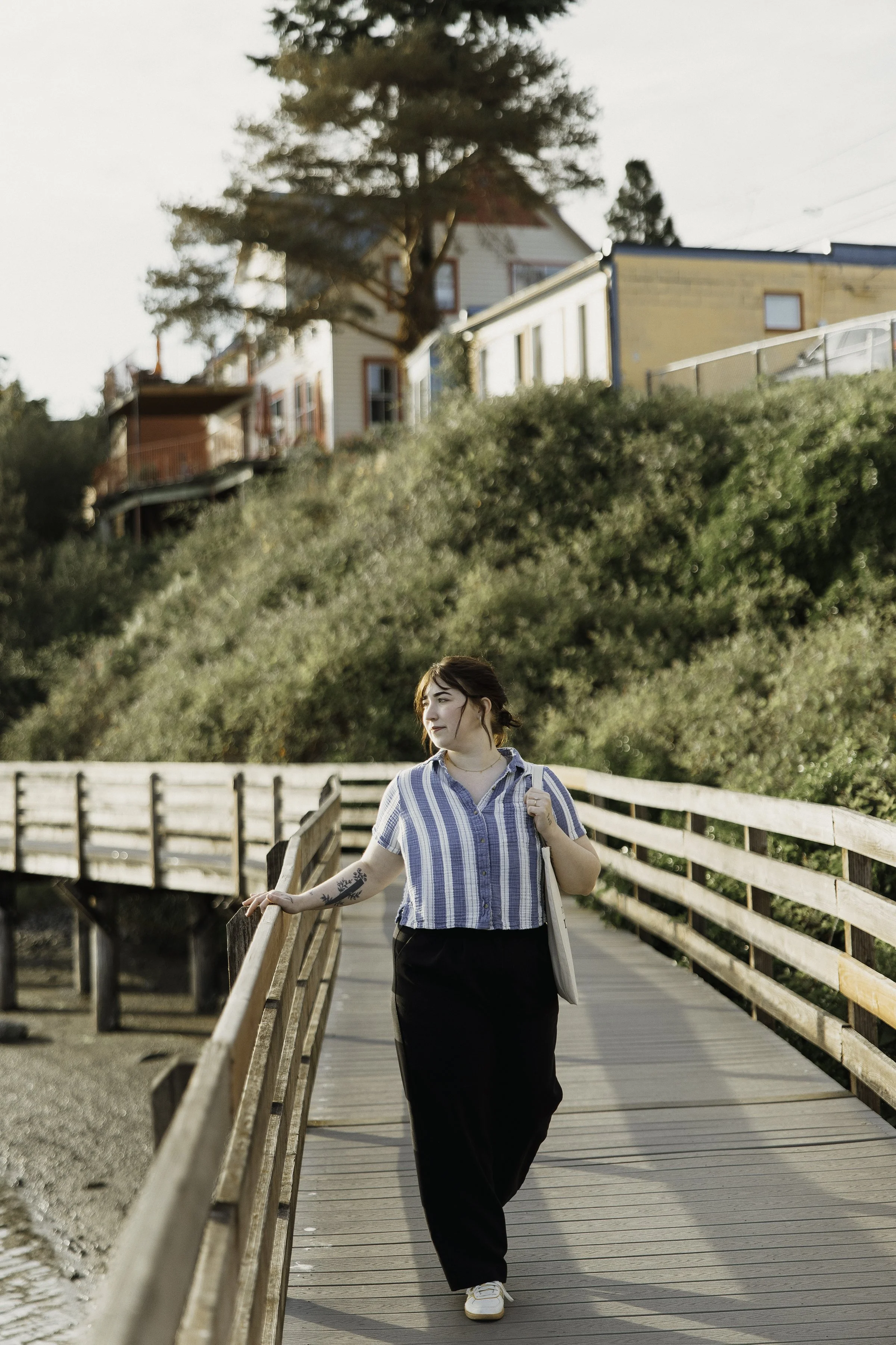 A woman holding a tote bag walking on a wooden bridge in a residential area with houses and trees in the background.