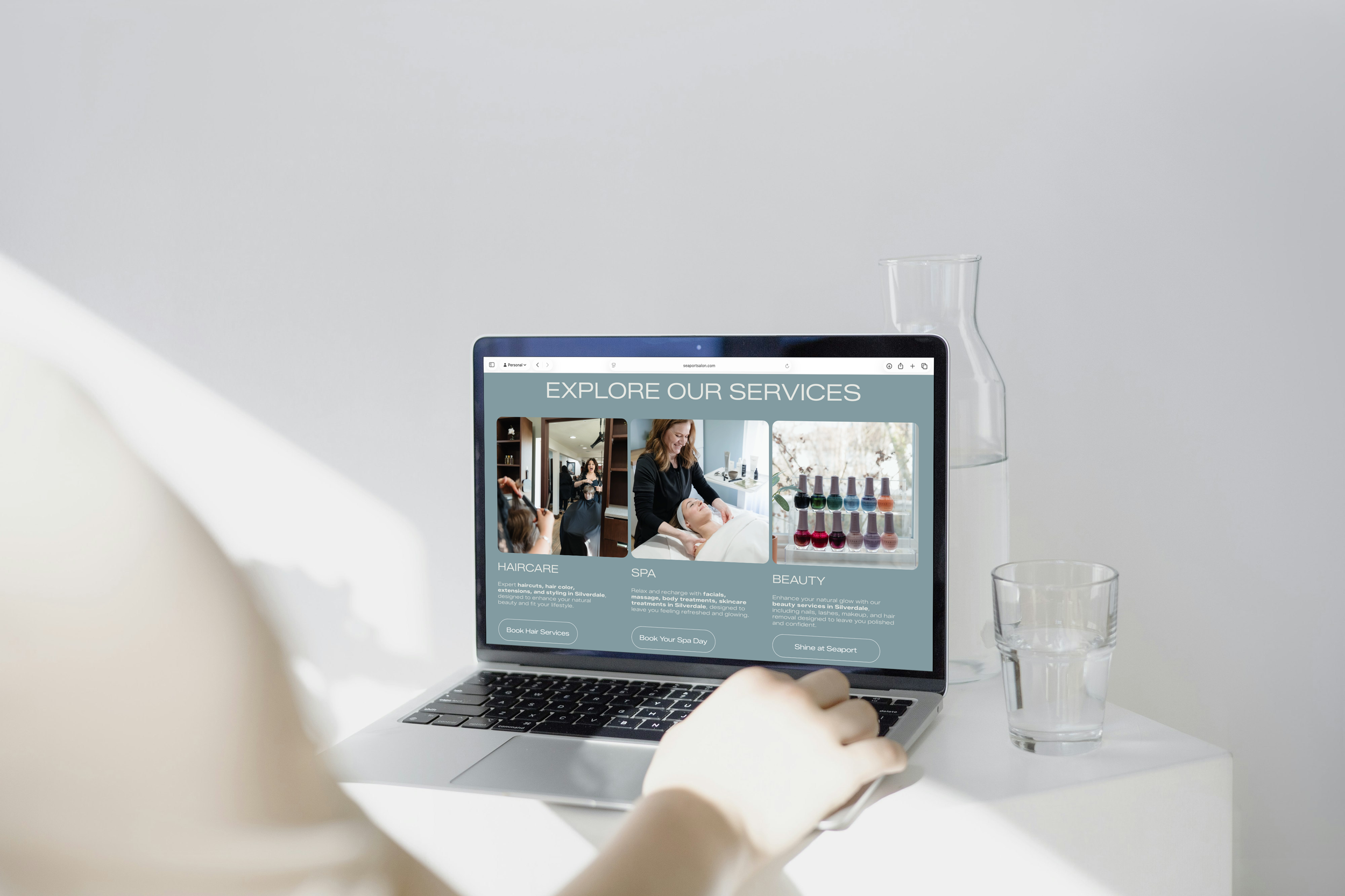 Laptop on a white table displaying an 'Explore Our Services' page for a spa, with options for Haircare, Spa, and Beauty services, next to a drinking glass and a tall glass vase.