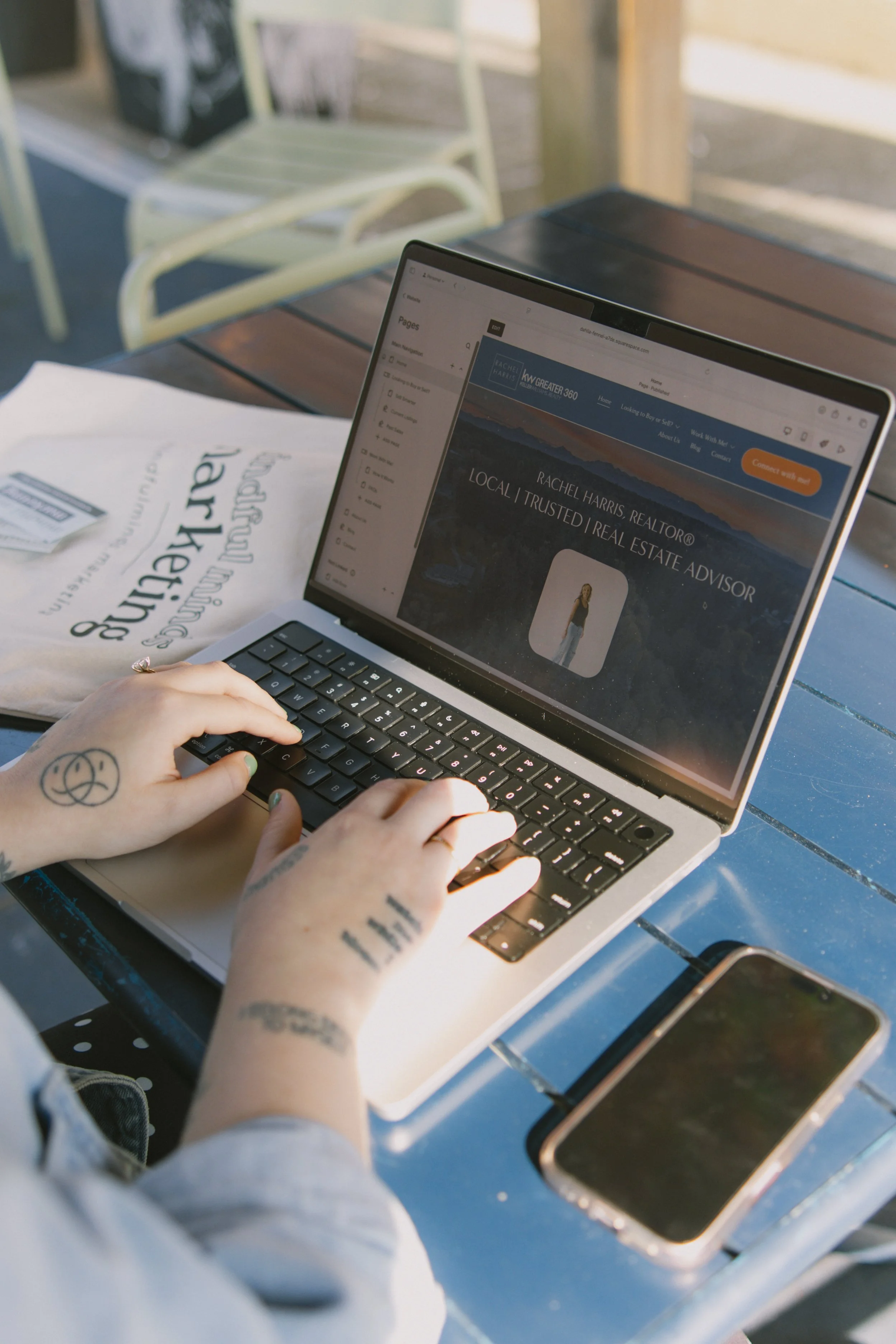 Person with tattoos on both arms using a laptop at a blue outdoor table, with a smartphone nearby and a paper bag on the table, in an outdoor setting with chairs.