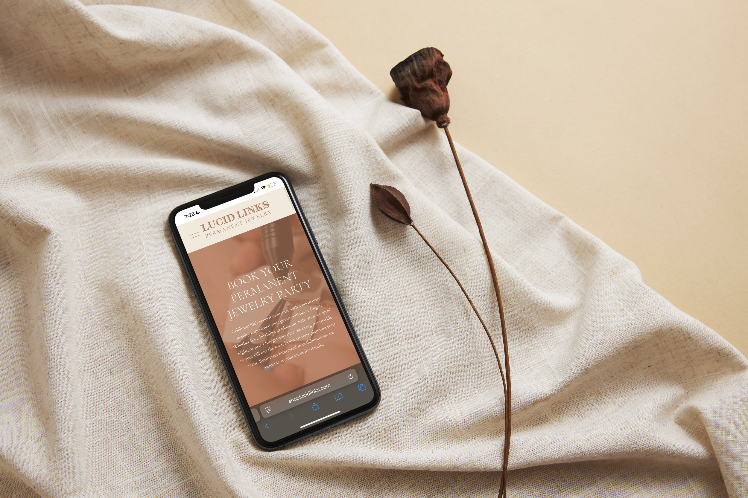 Smartphone displaying website for Lucid Links, a jewelry company, placed on a cream-colored fabric next to two dried brown flowers. The phone screen shows a promotion to book a permanent jewelry party.