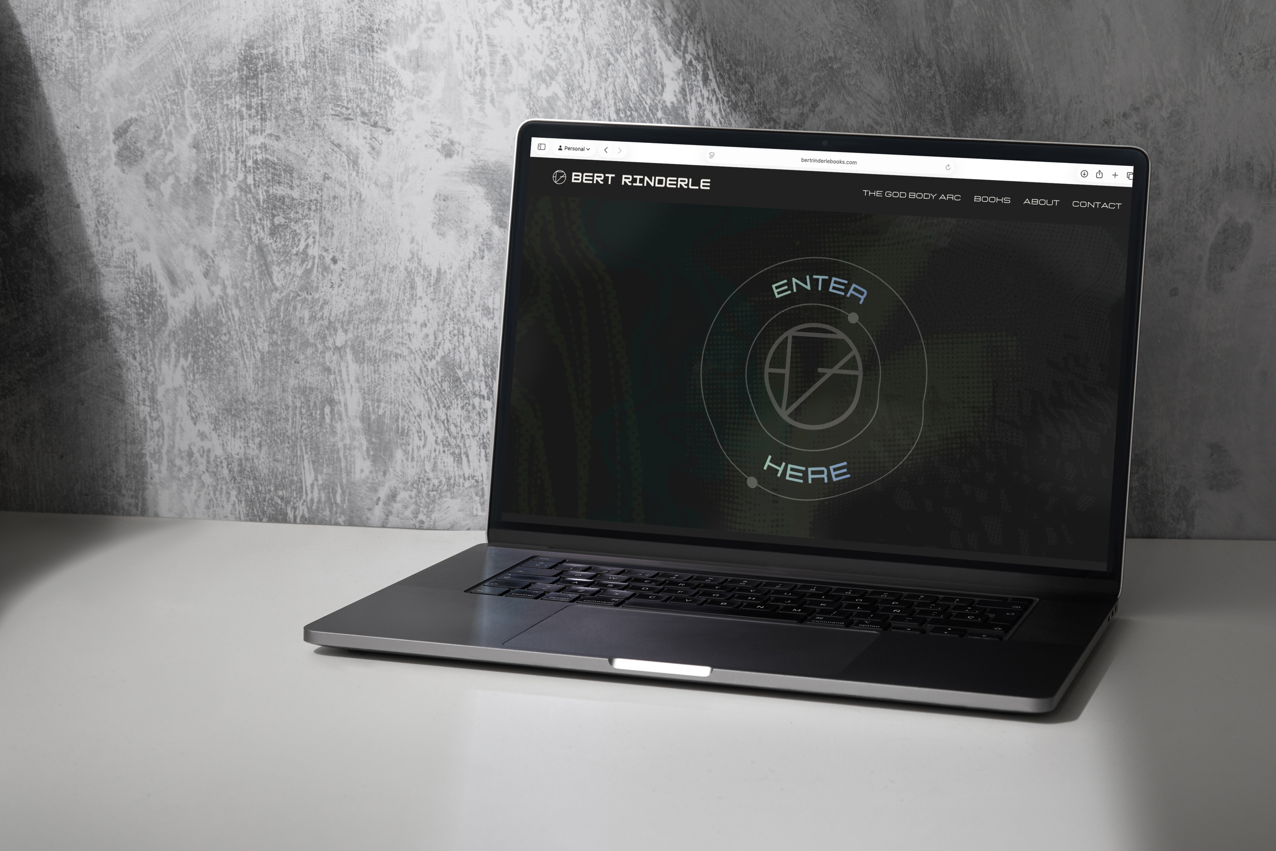 A laptop on a white desk with a website open displaying the name Bert Rinderle and the words 'Enter Here' on a dark background, against a textured gray wall.