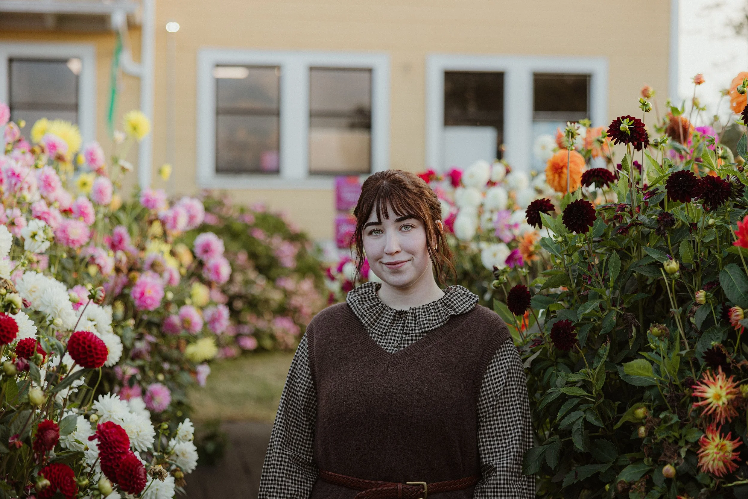 A young woman with reddish-brown hair and bangs, wearing a checked shirt and a sleeveless brown sweater, standing amid colorful blooming flowers outdoors with a house in the background.