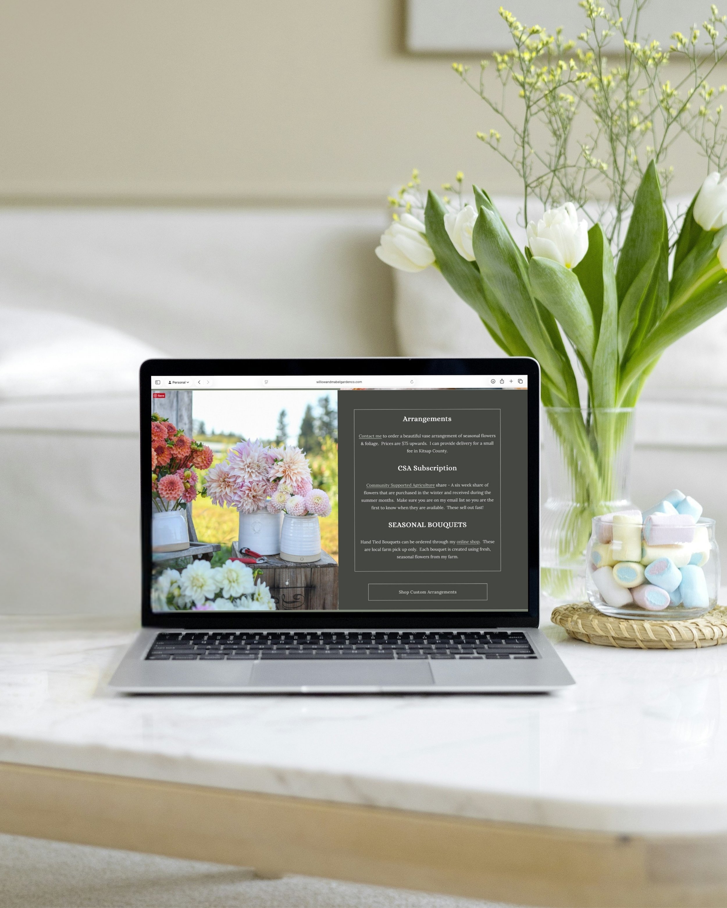 A laptop on a white table displaying a website for flower arrangements, with a vase of tulips and a jar of marshmallows nearby.