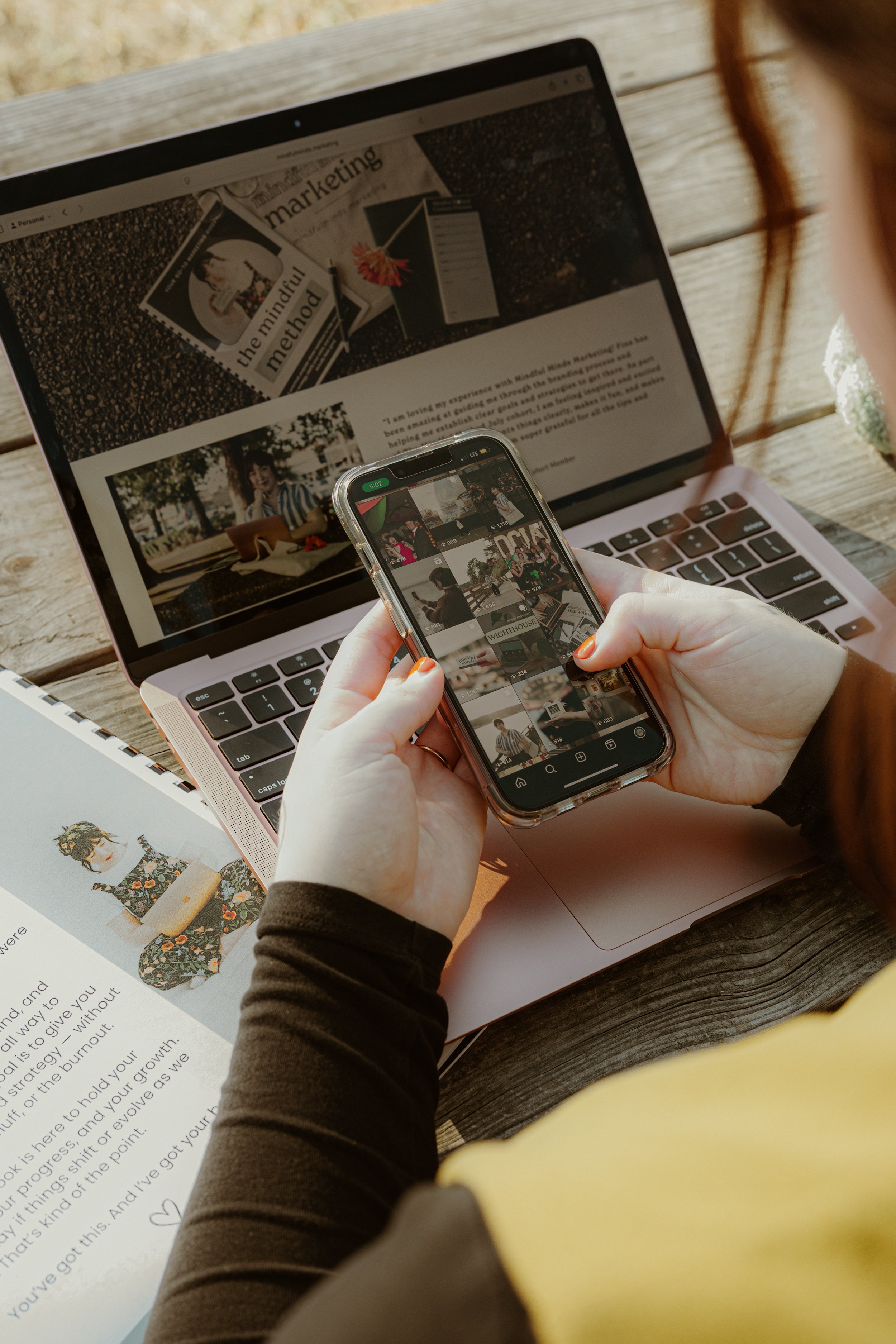 A person taking a photo of a laptop and a magazine with a smartphone outside. The laptop shows a webpage with images and text, and the magazine has a picture of a woman sitting outdoors.