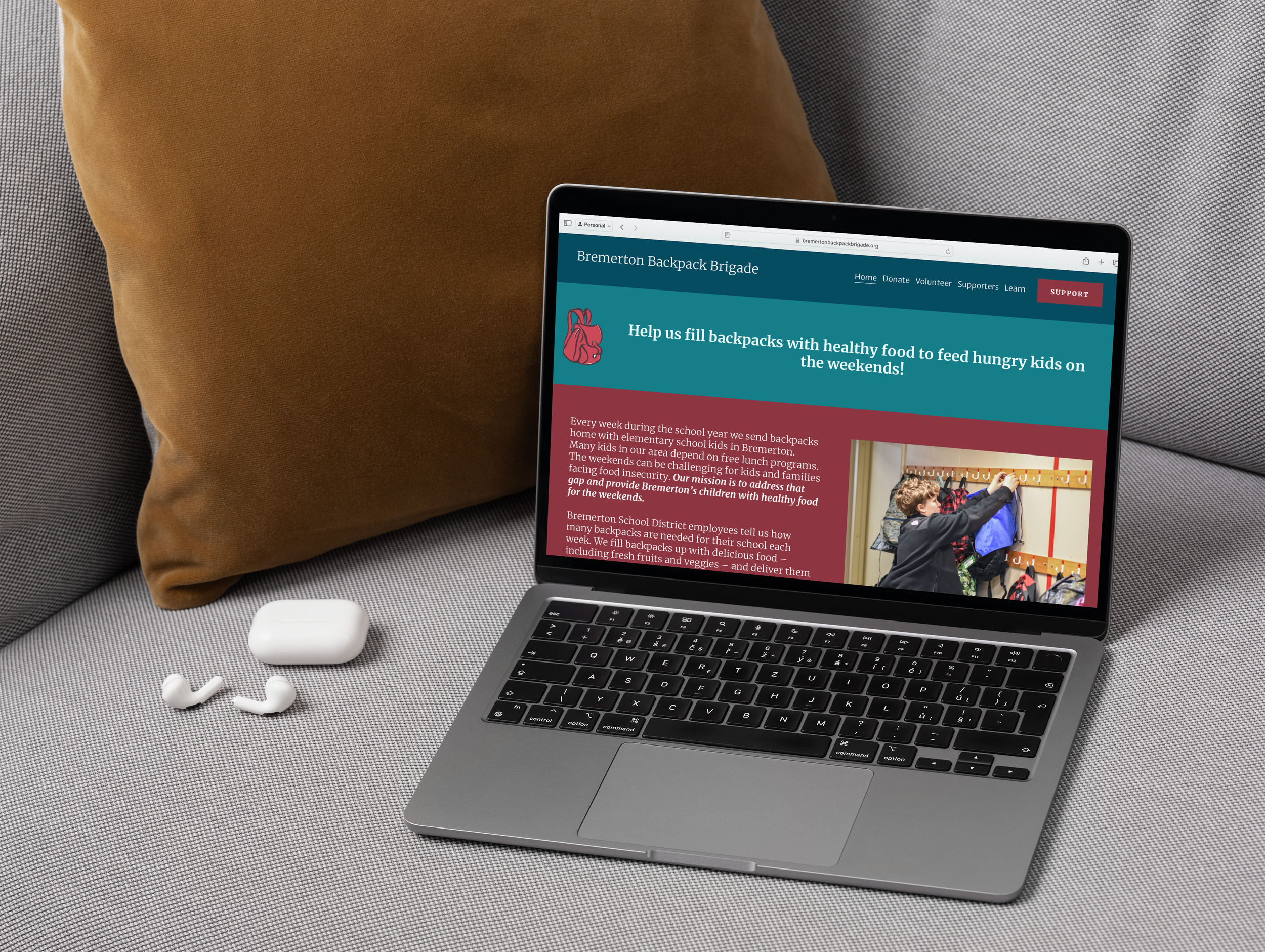 A laptop displaying the Bremerton Backpack Brigade website, placed on a light-colored textured surface with a brown cushion behind it, along with a pair of white earbuds and their case nearby.