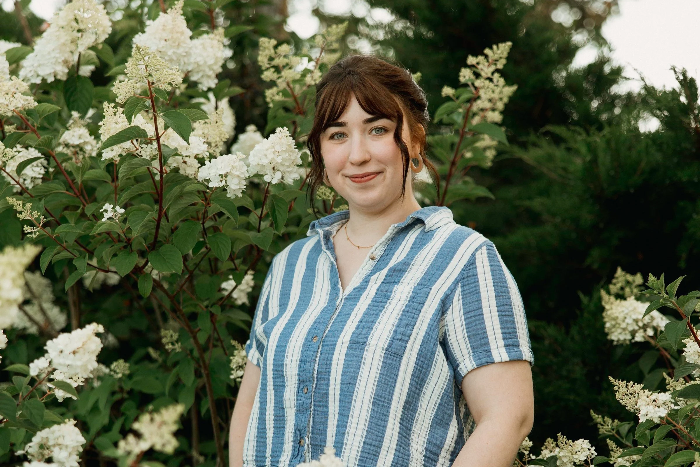 A young woman with brown hair and blue eyes is smiling outdoors amid white flowering shrubs, wearing a blue and white striped shirt.