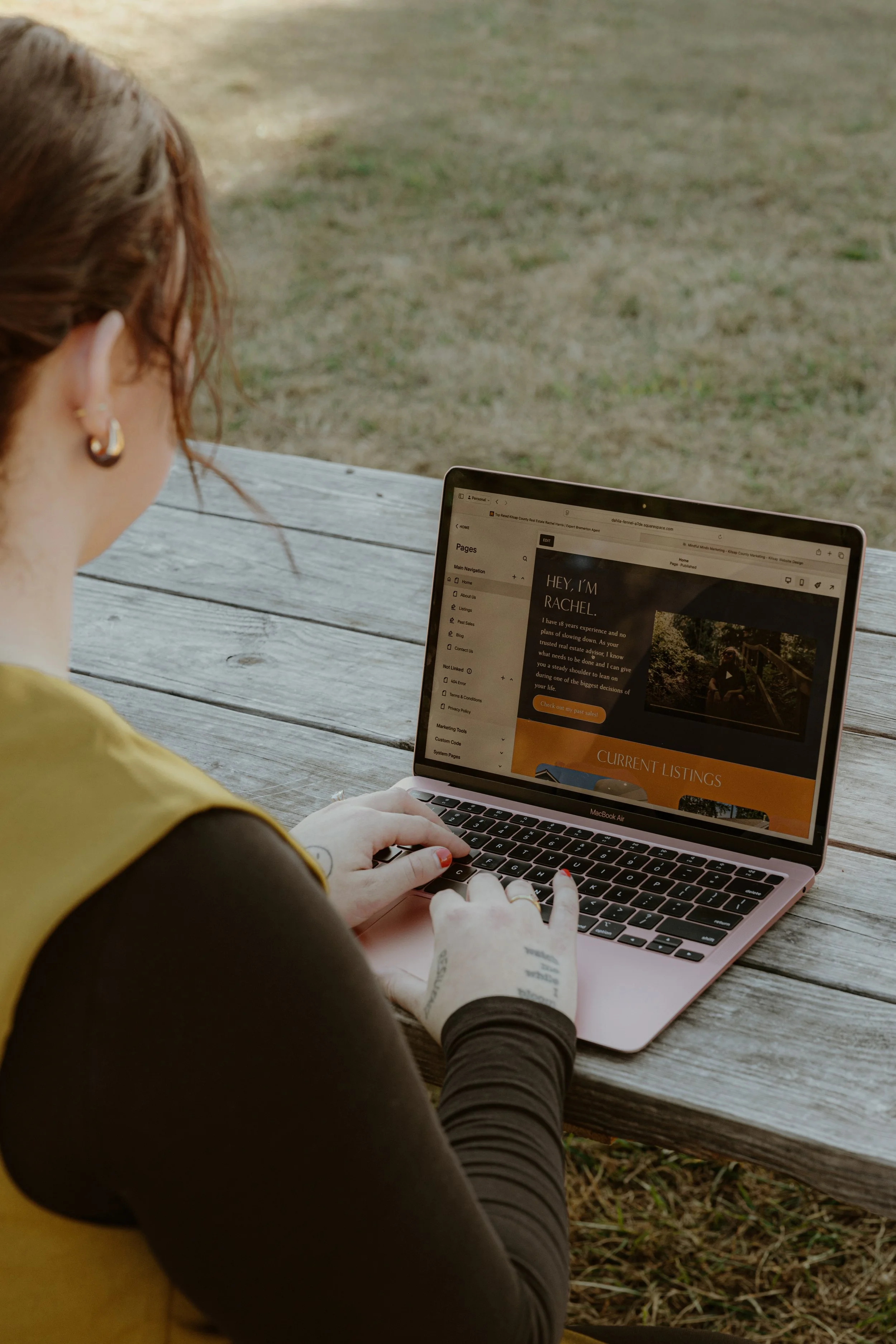 A person wearing a striped shirt typing on a rose gold laptop on a red outdoor table, with a smartphone, a green notebook, and glasses nearby.