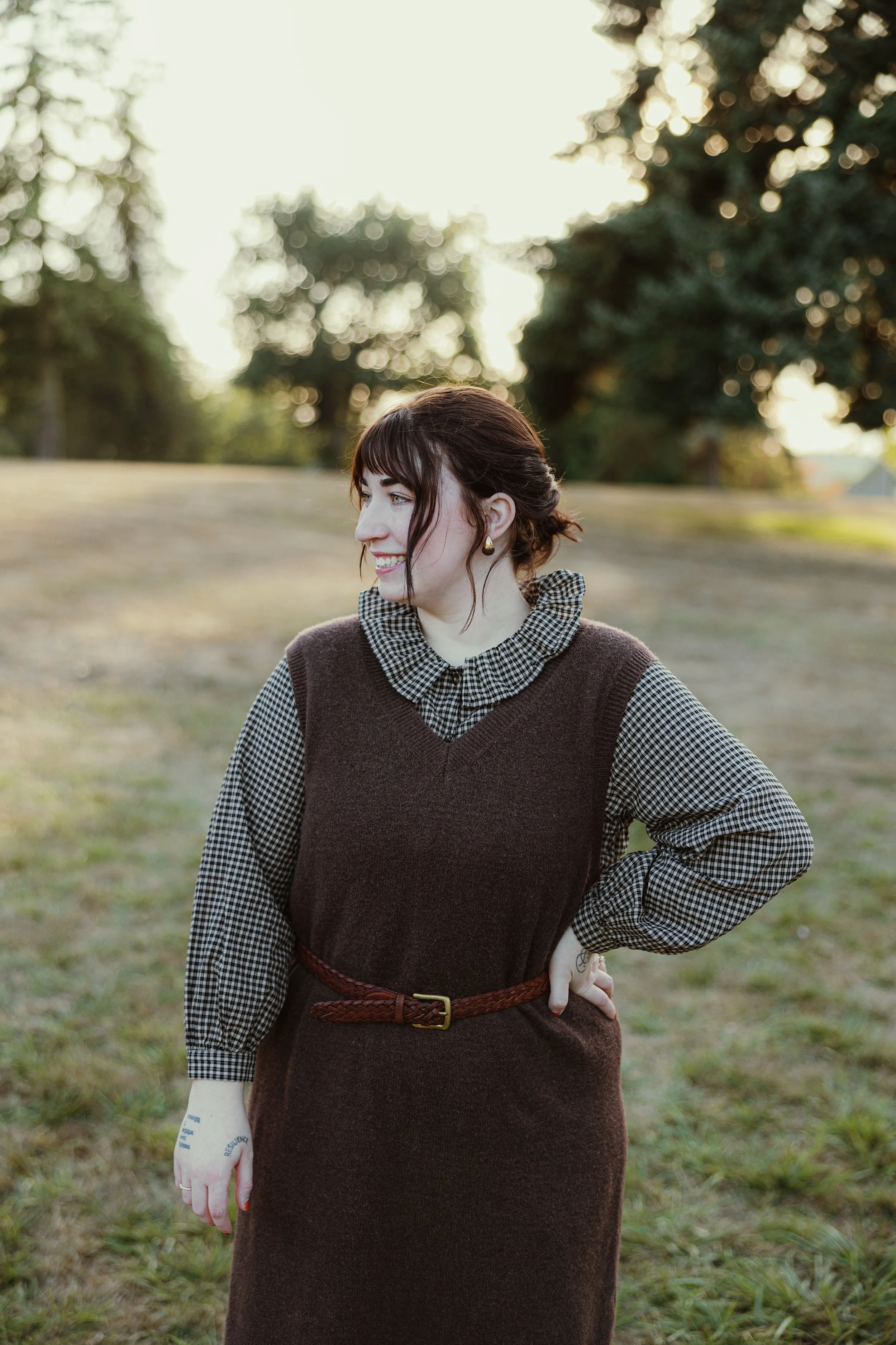 A young woman with brown hair and bangs, wearing a brown sleeveless dress over a checkered shirt, standing outdoors on a grassy field with trees in the background during sunset. She is smiling, posing with one hand on her hip.