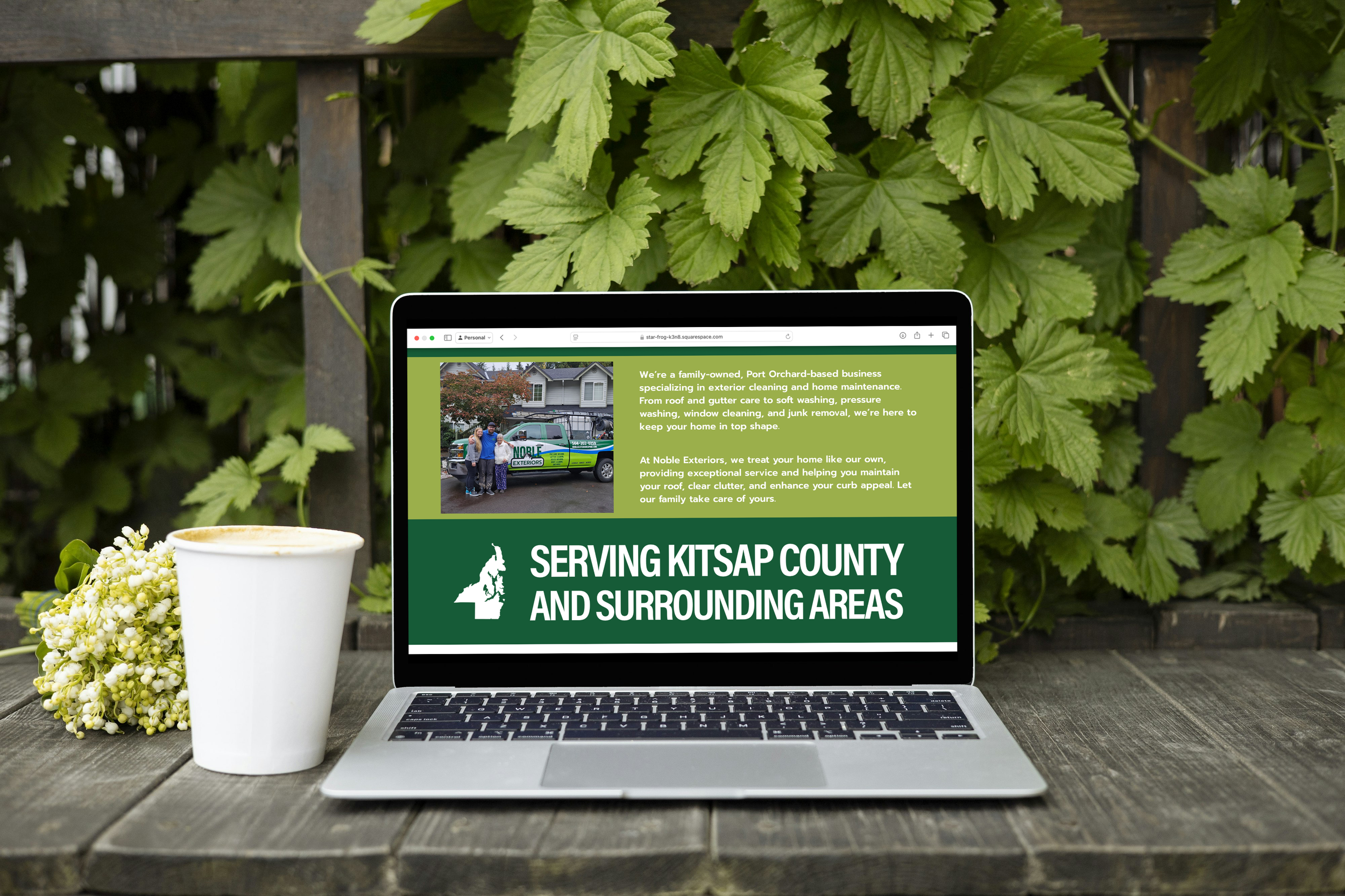 Laptop displaying a landscaping company's website in front of green plants, accompanied by a white flower bouquet and a white to-go coffee cup on a wooden surface.