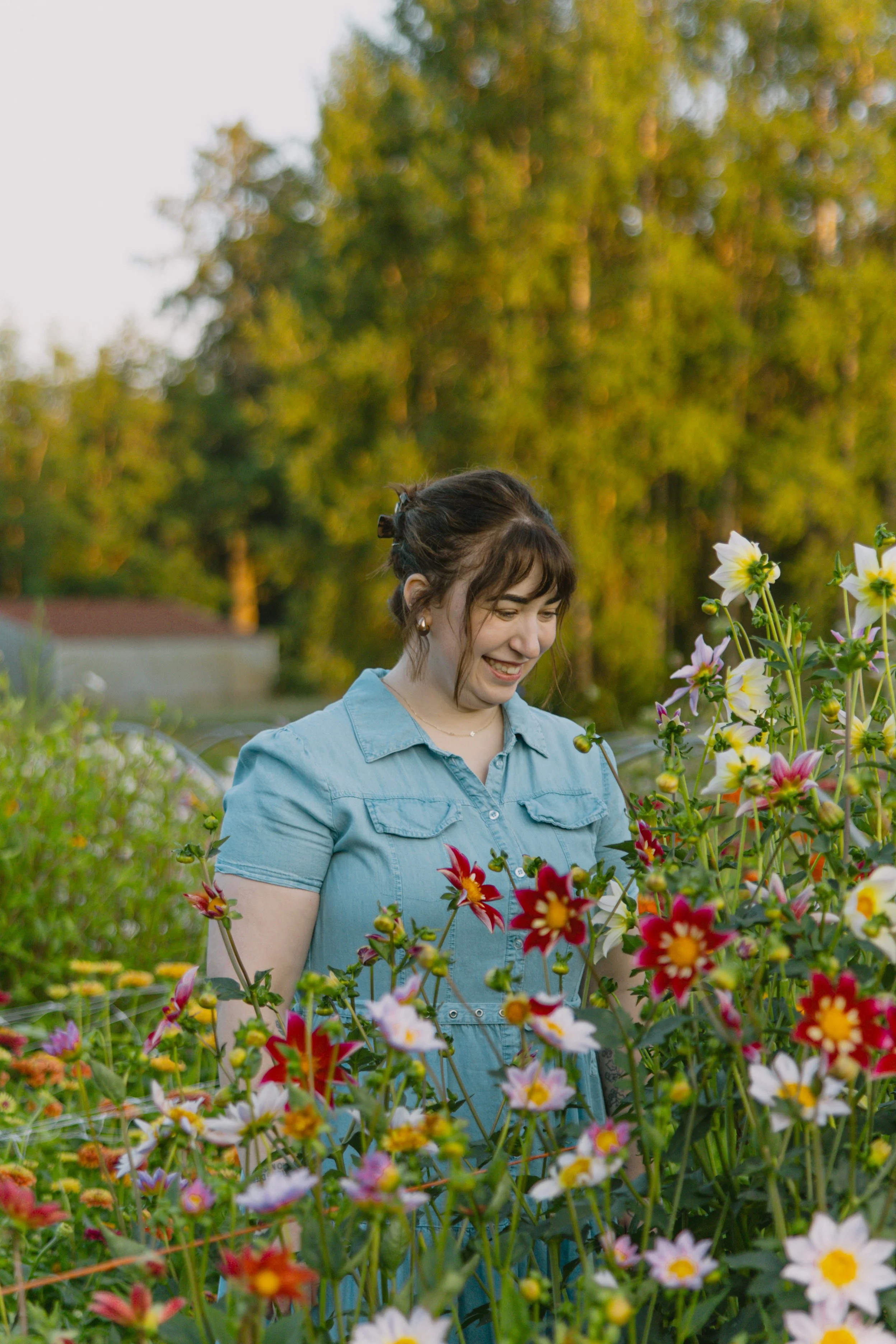 A smiling woman with dark hair in a bun, wearing a light blue button-up shirt, standing in a garden of colorful flowers with a background of trees and a clear sky.