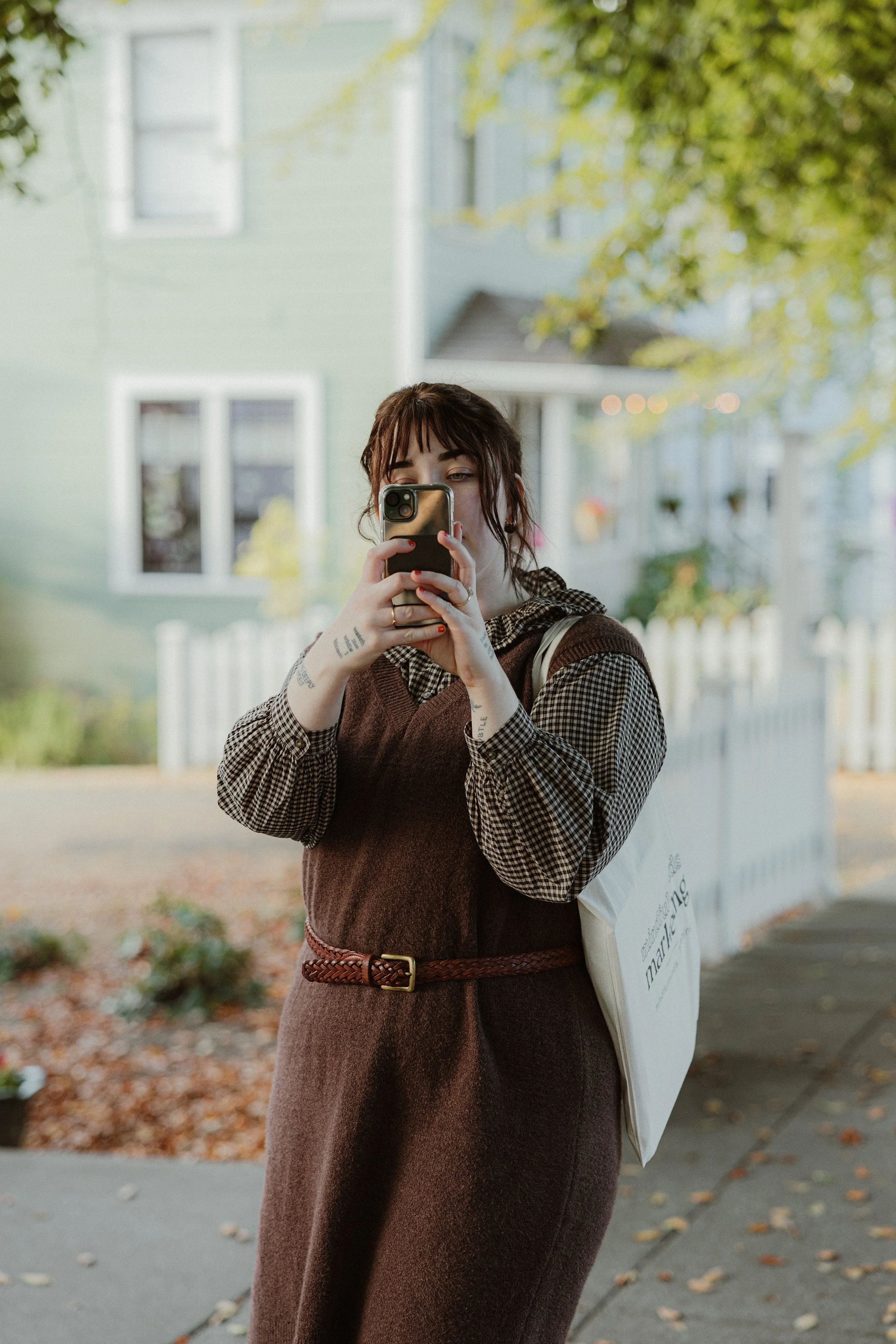A woman with brown hair and tattoos on her arms stands on a sidewalk outside, taking a photo with her smartphone. She is wearing a brown dress over a checked long-sleeve shirt, with a belt, and carries a white tote bag. There are green trees and a pastel-colored house in the background.
