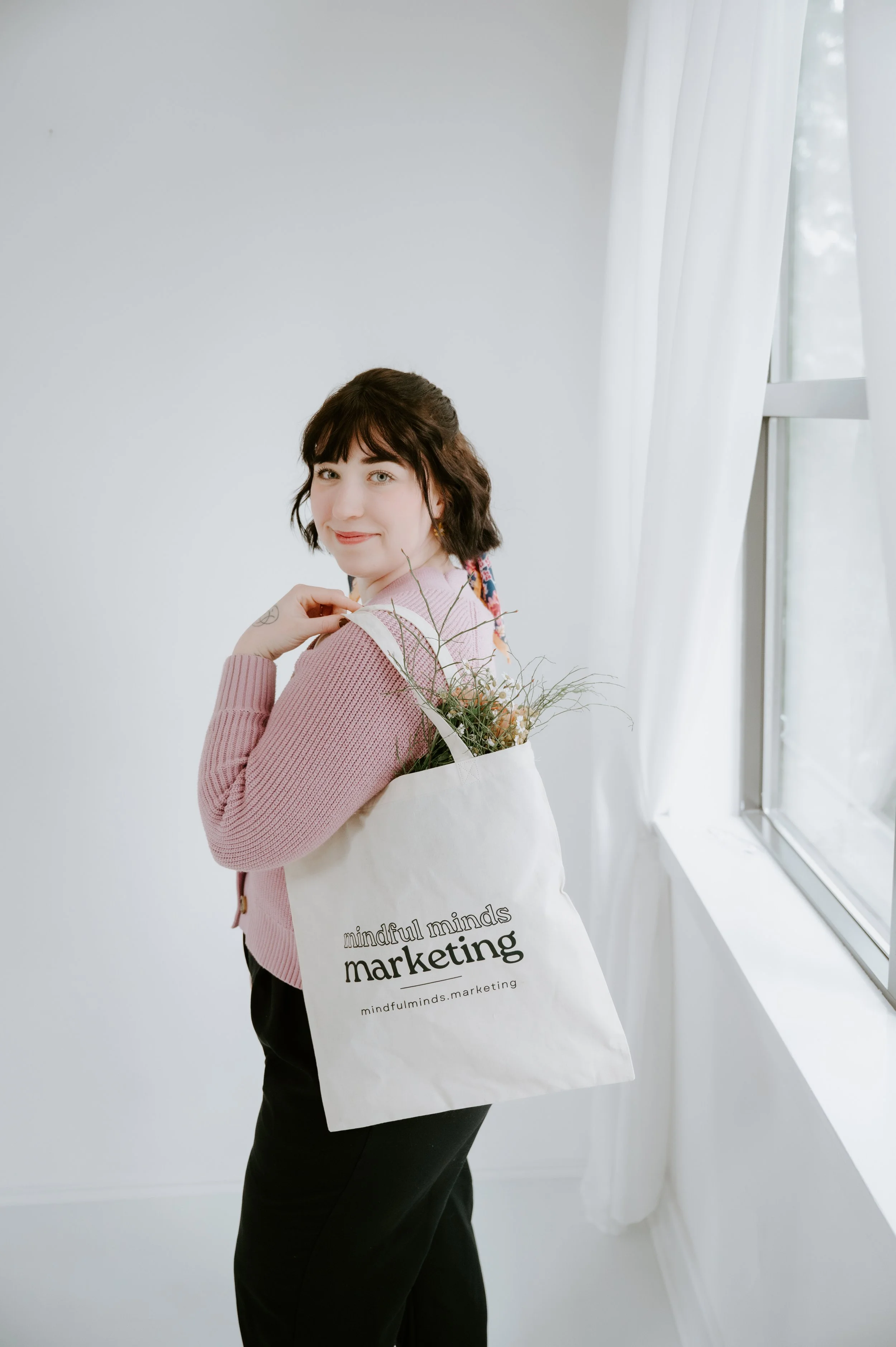 A woman standing indoors near a window holding a tote bag with flowers, wearing a pink sweater and black pants, smiling at the camera.