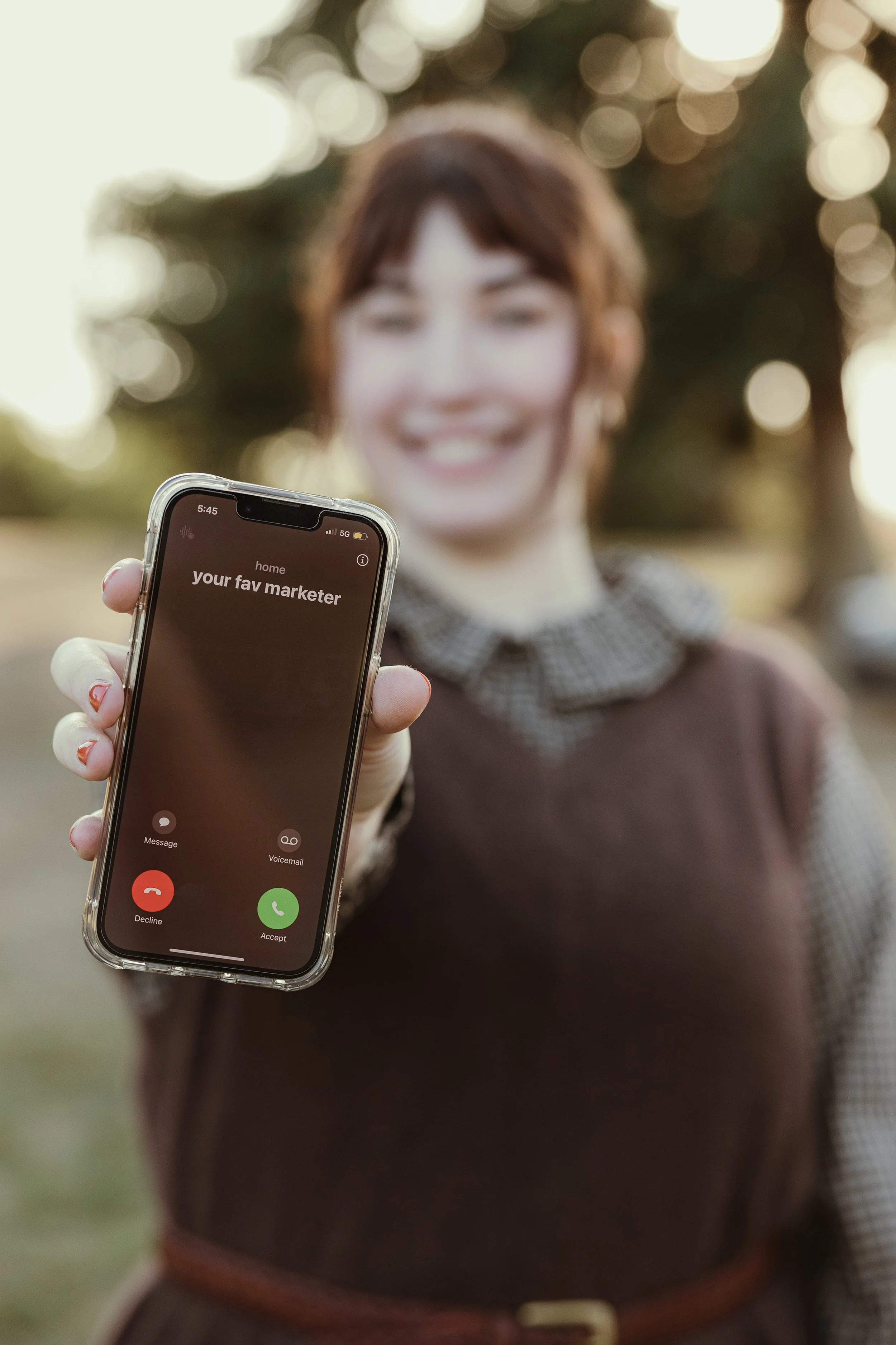 A woman with glasses and a striped shirt working on a laptop at an outdoor cafe, with orange and blue flags and trees in the background.