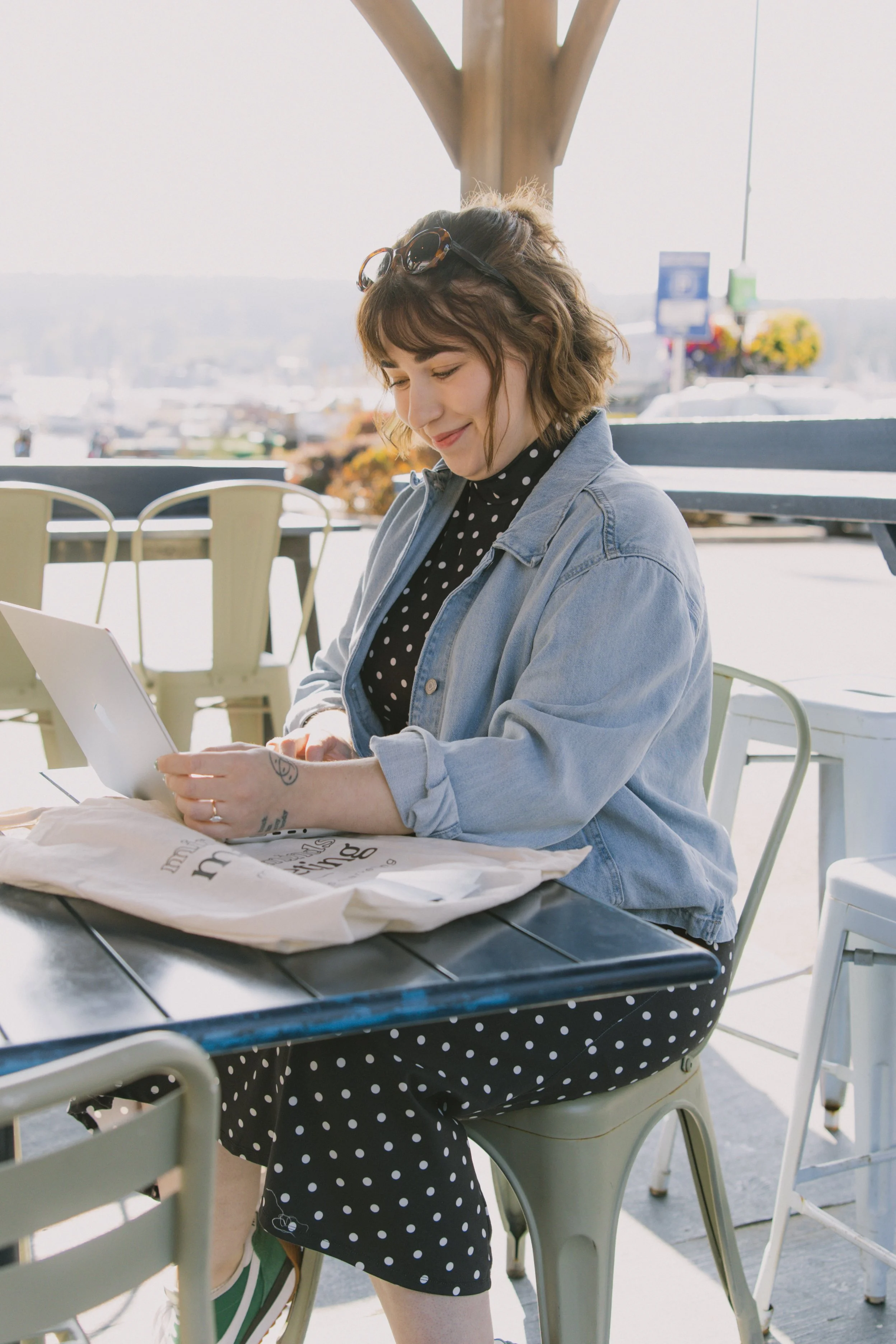 A woman with glasses and a striped shirt working on a laptop at an outdoor cafe, with orange and blue flags and trees in the background.