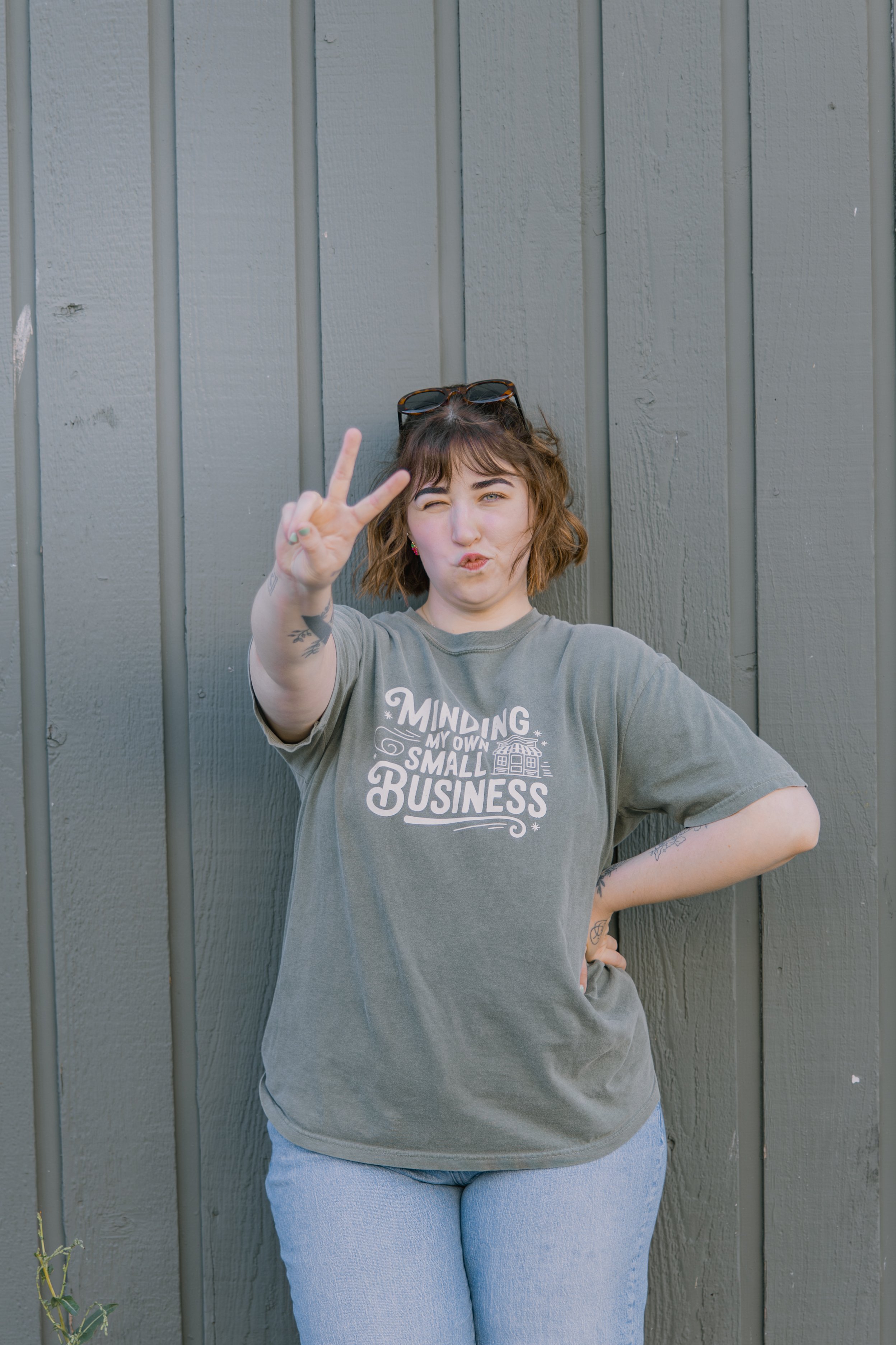 A woman with short, wavy brown hair standing against a gray wooden fence, making a peace sign with her right hand and winking at the camera, wearing sunglasses on her head, a gray t-shirt with white text, and light blue jeans.