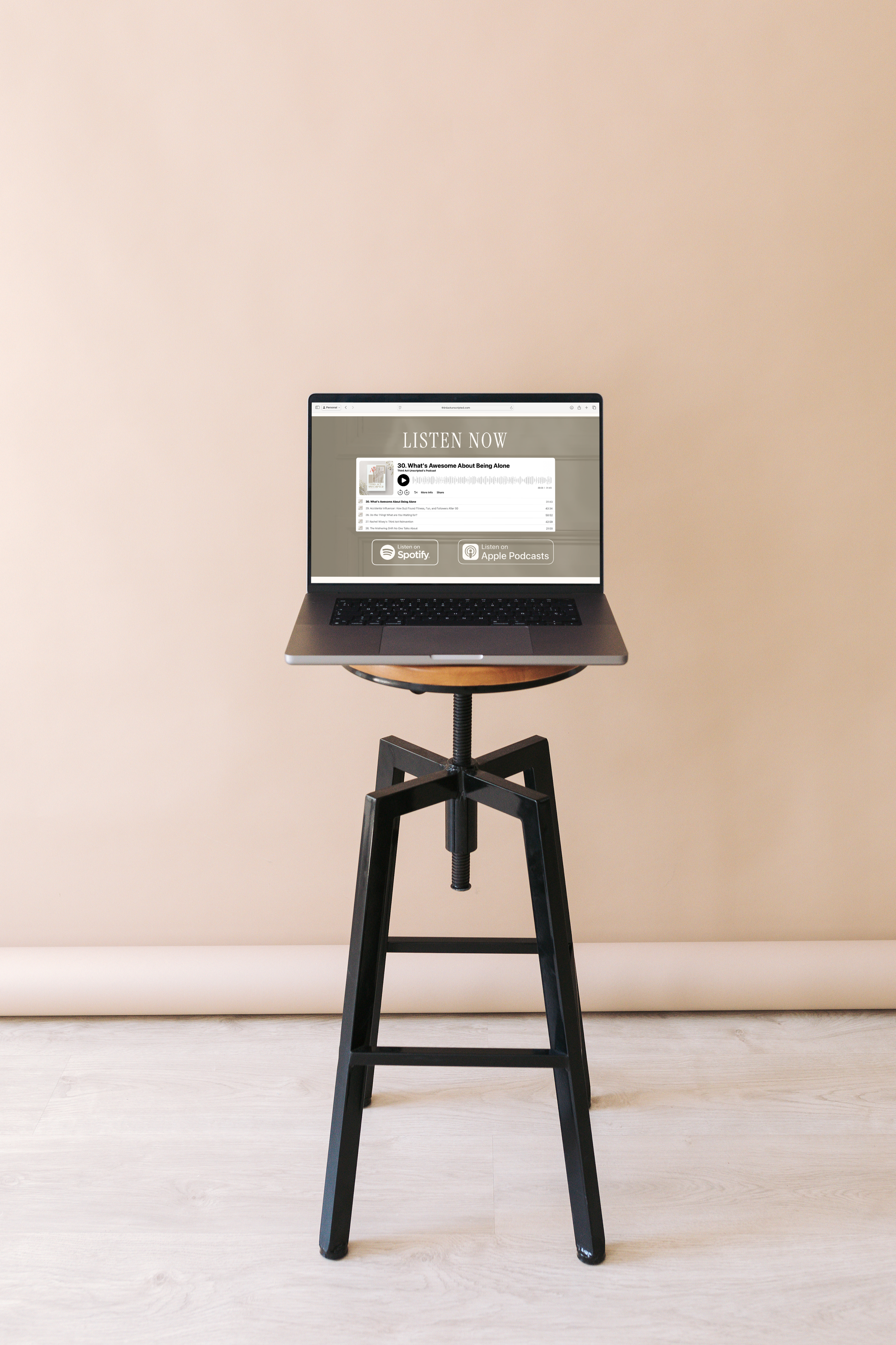 Laptop on a tripod stand displaying a music player with options to listen via Spotify or Apple Podcasts against a plain background.