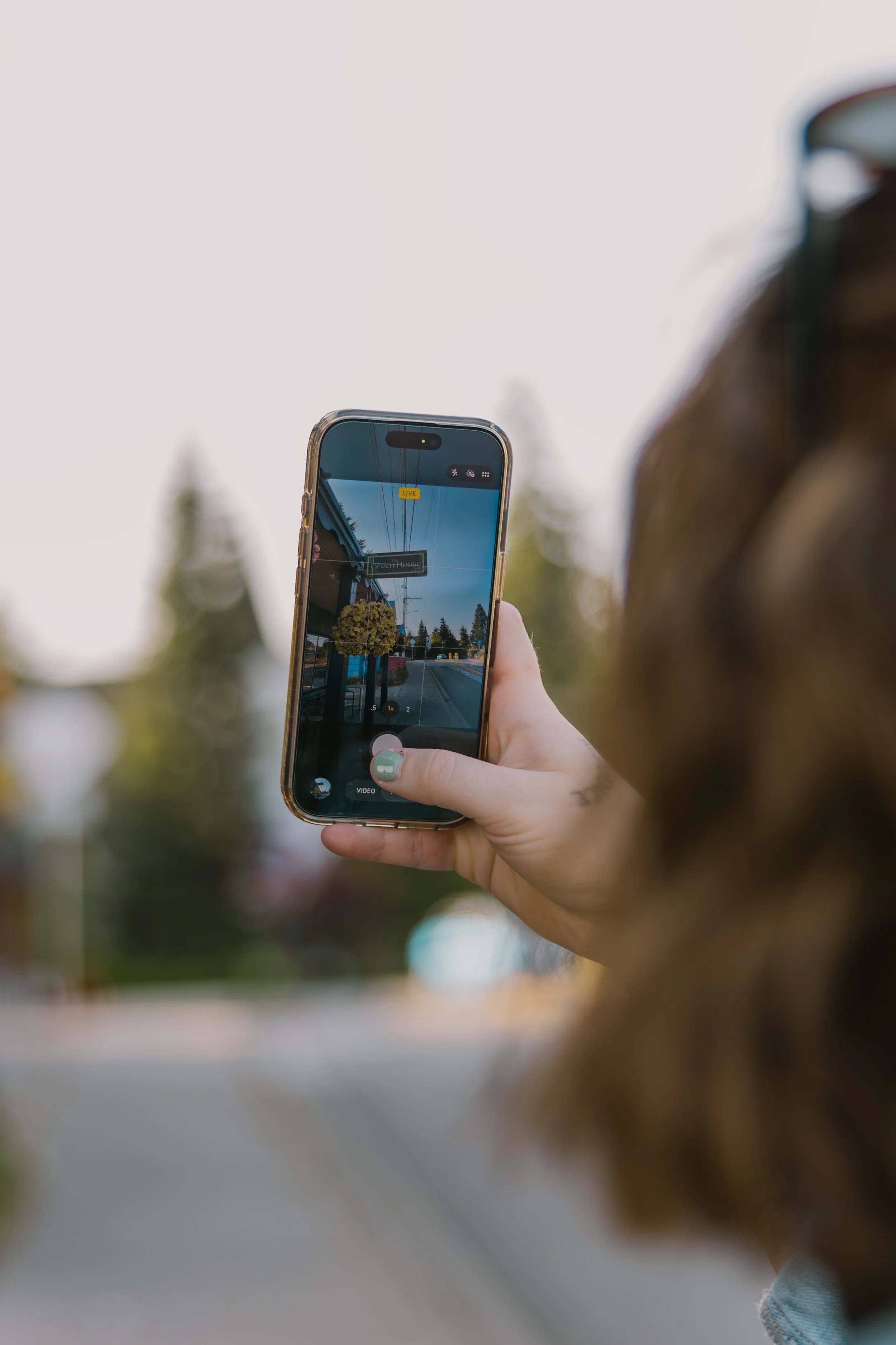 Person holding a smartphone in portrait orientation, capturing a photo of a street with buildings, trees, and a sign that reads 'The Little House' in the background, outdoors during daytime.