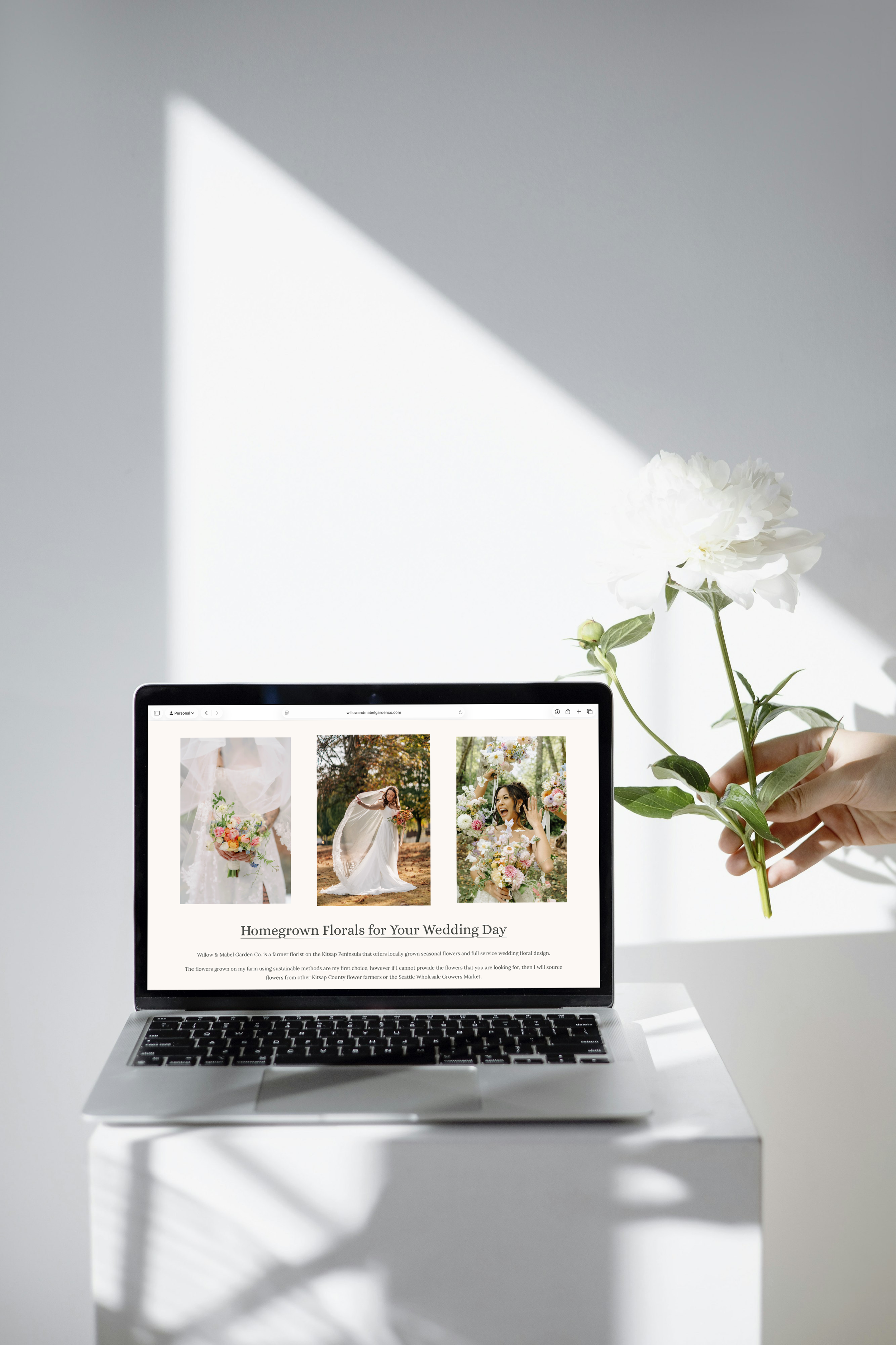 Laptop displaying images of homegrown floral arrangements for a wedding, with a person's hand holding a white flower beside the laptop.