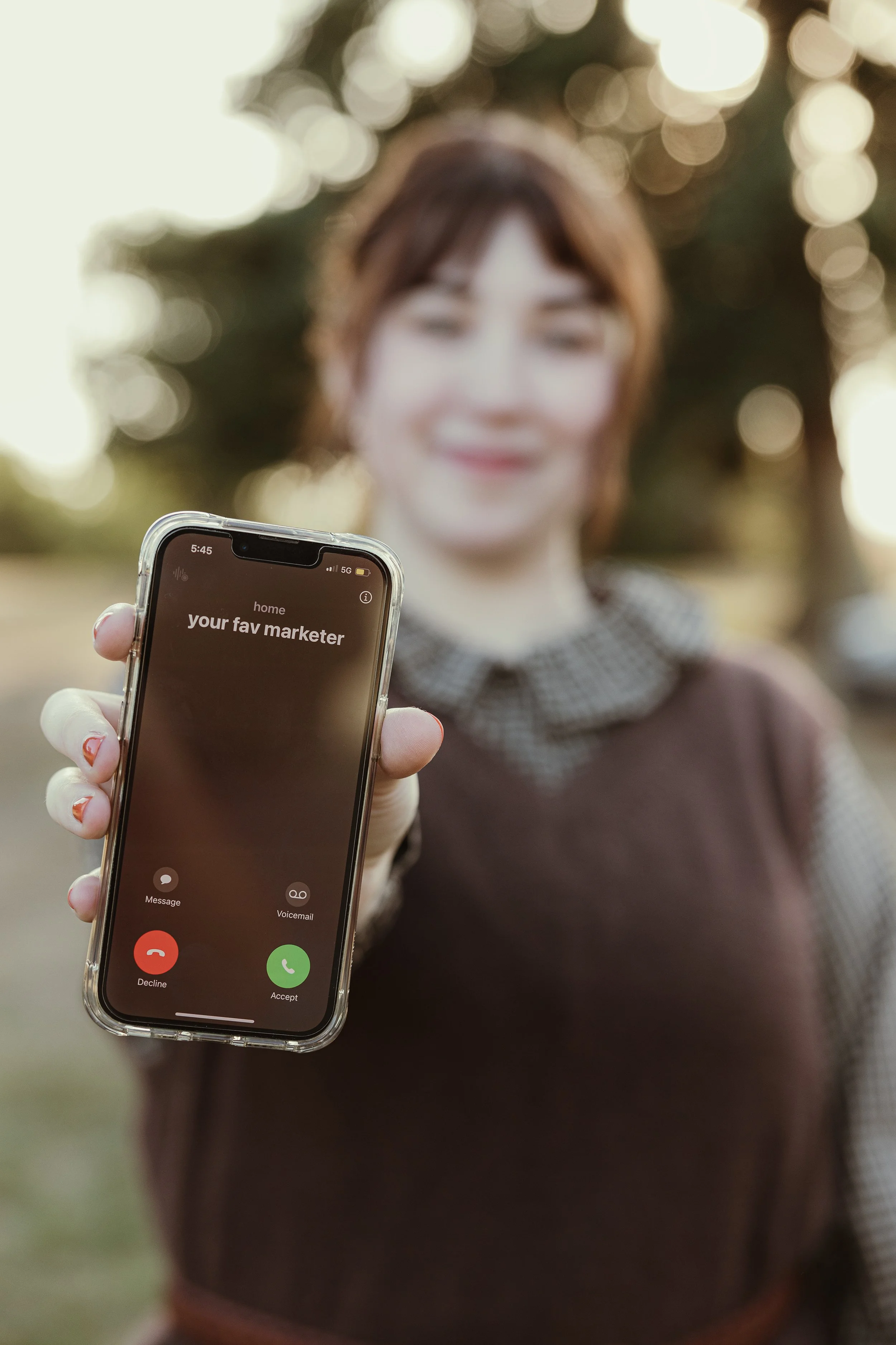 A woman holding a smartphone showing an incoming call from 'your fav marketer' outdoors with blurred trees in the background.