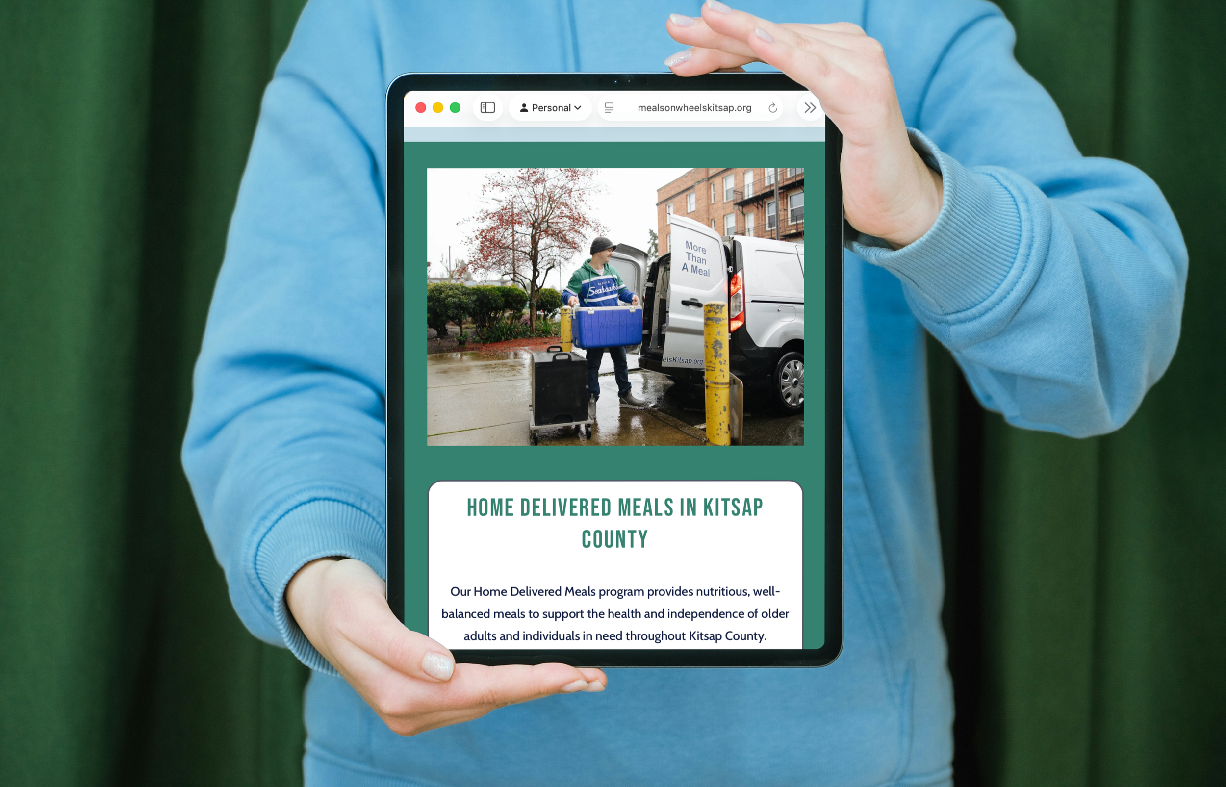 Person holding a tablet displaying a website for Meals on Wheels Kitsap County, showing a delivery person unloading meals from a van on a rainy day.