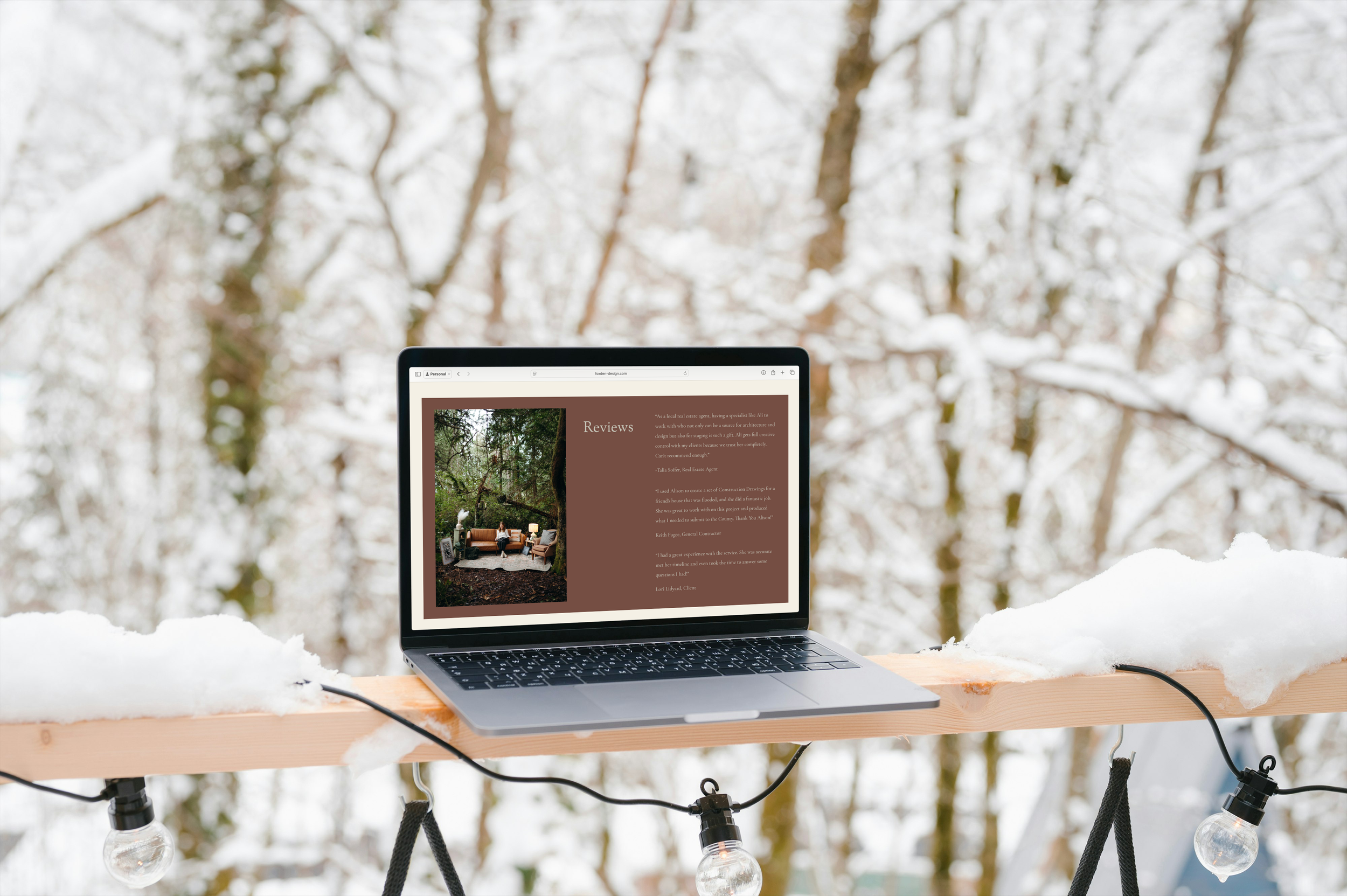 A laptop displaying a review webpage with outdoor furniture scene on a wooden railing, outdoors in snowy forest with string lights.