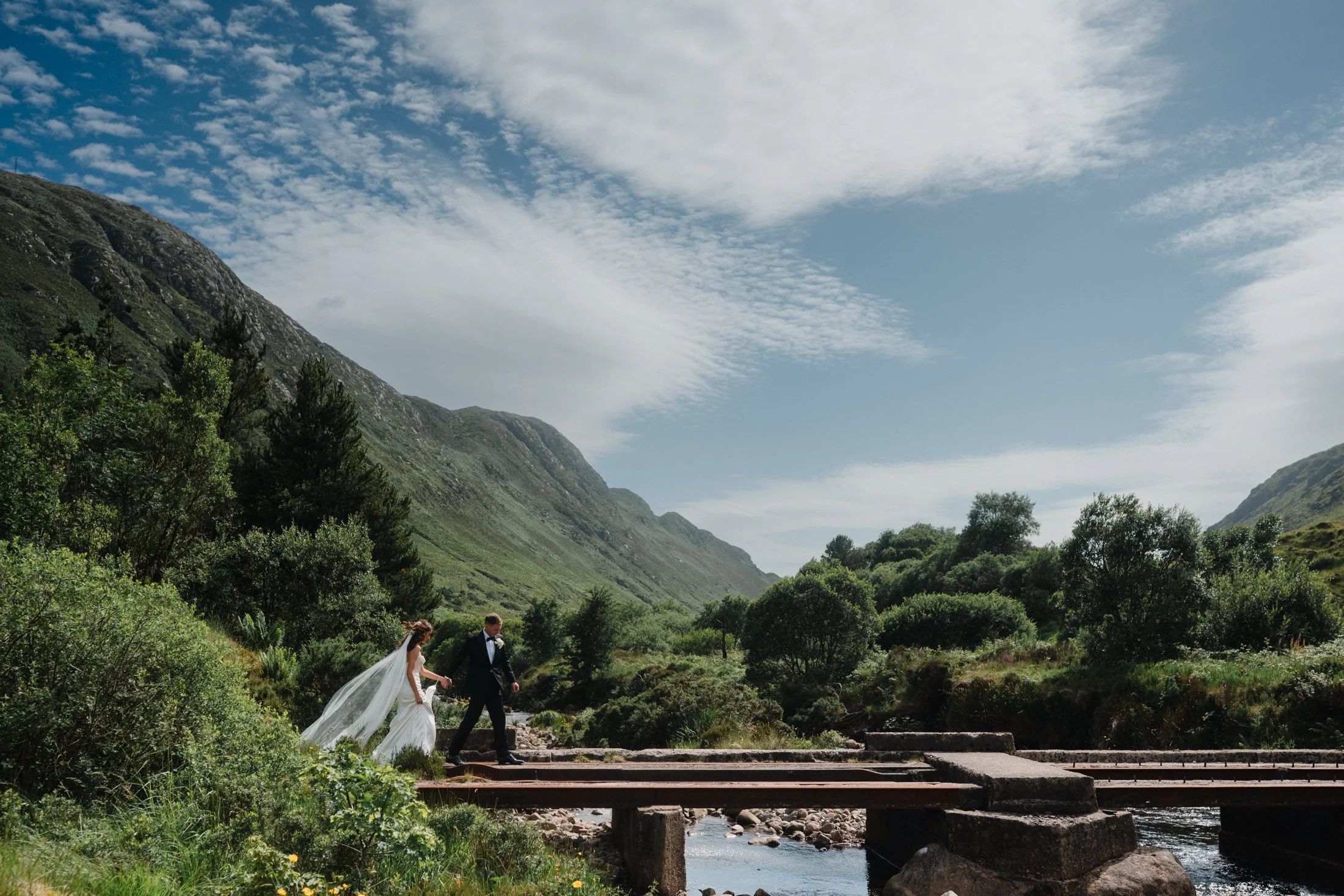 A bride and groom walking across a small rustic wooden bridge in donegal hills over a stream in a lush green valley with mountains and partly cloudy sky in the background.
