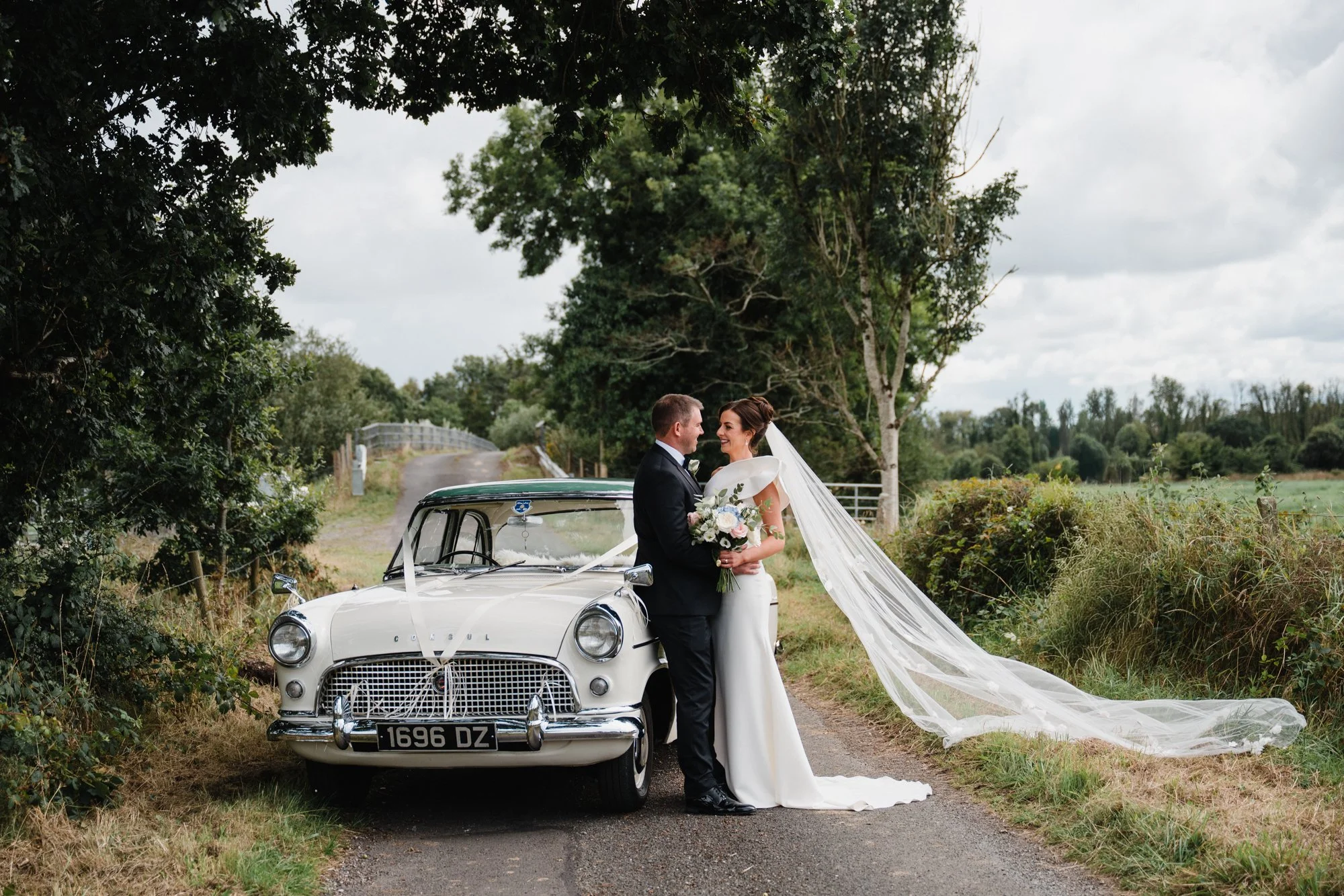 A bride and groom standing together in front of a vintage white car decorated with white ribbons. This Irish bride is holding a bouquet, and they are smiling at each other outdoors on a rural path with trees and cloudy sky in the background.