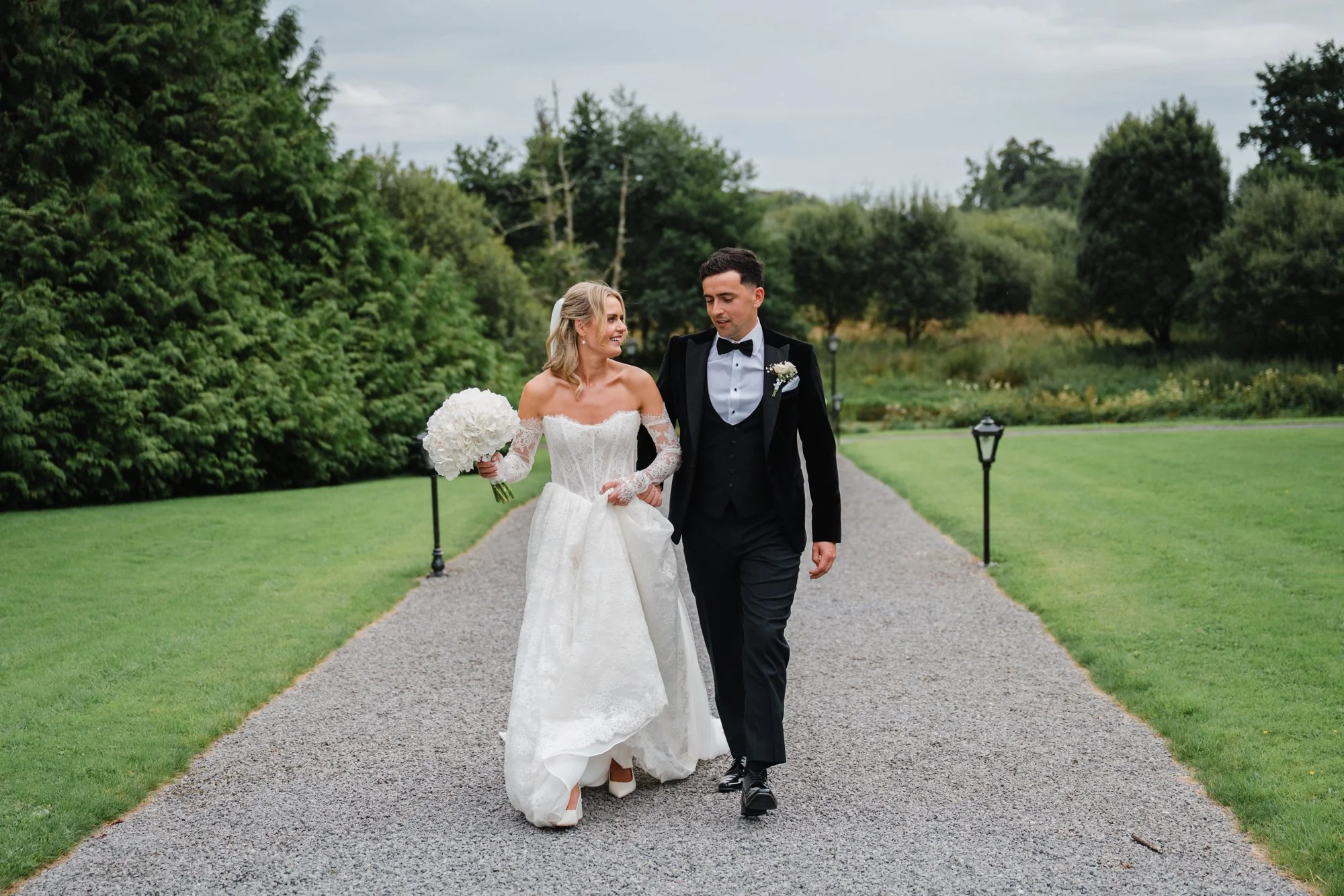 A bride and groom walking down a gravel path on their wedding day outdoors. The bride is smiling and holding a bouquet of white flowers, wearing an off-shoulder wedding dress. The groom is dressed in a black tuxedo with a bow tie and is looking at th