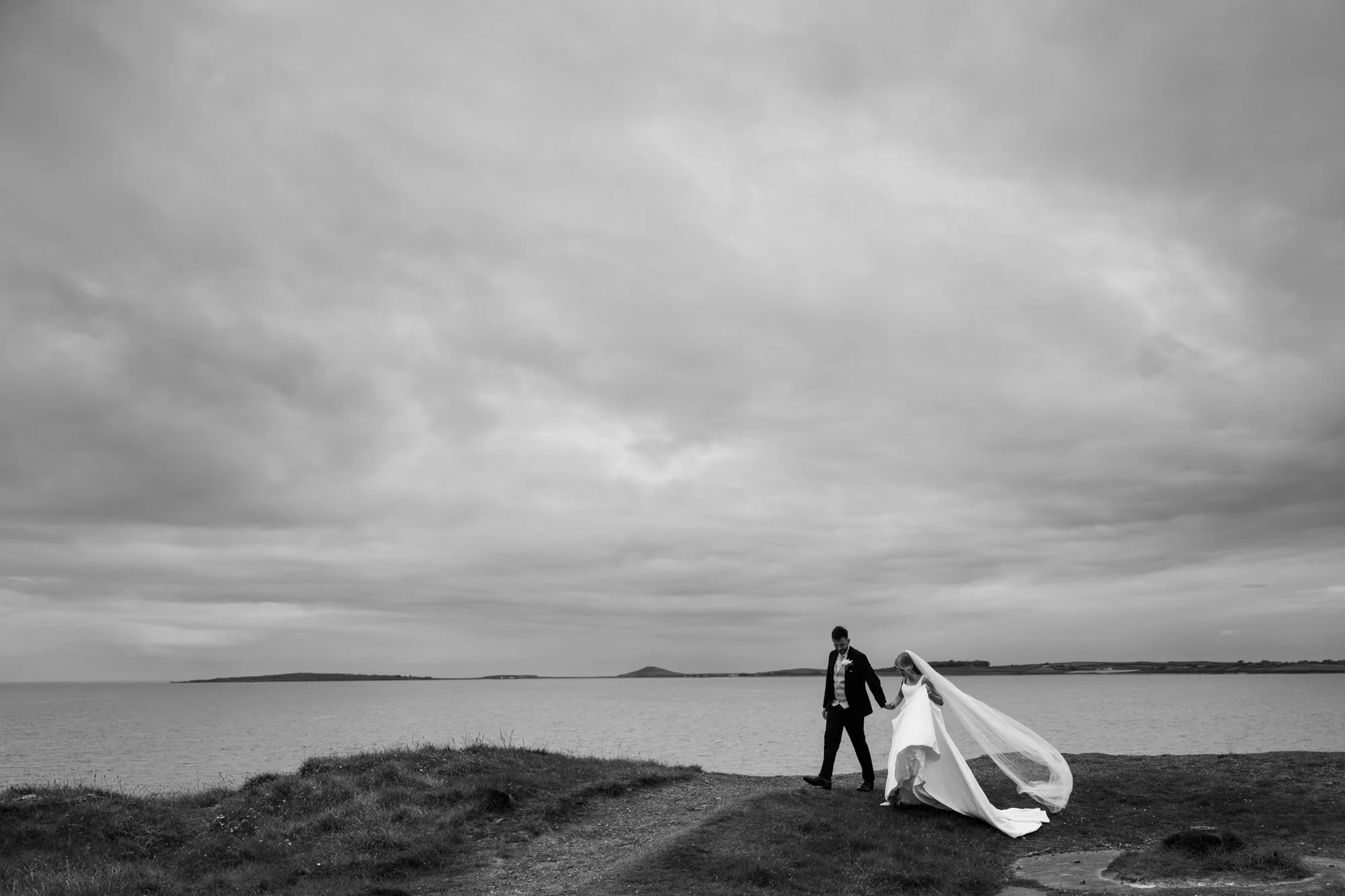 Black and white photograph of a bride and groom walking along the shoreline in Sligo  on a cloudy day, holding hands with the bride's long veil flowing behind them.