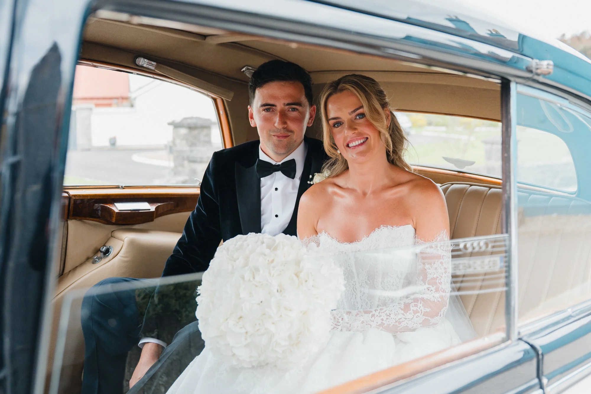 A bride and groom sitting inside a classic car, smiling, dressed in wedding attire, with the bride in a lace gown and the groom in a tuxedo, with the car window framing them.