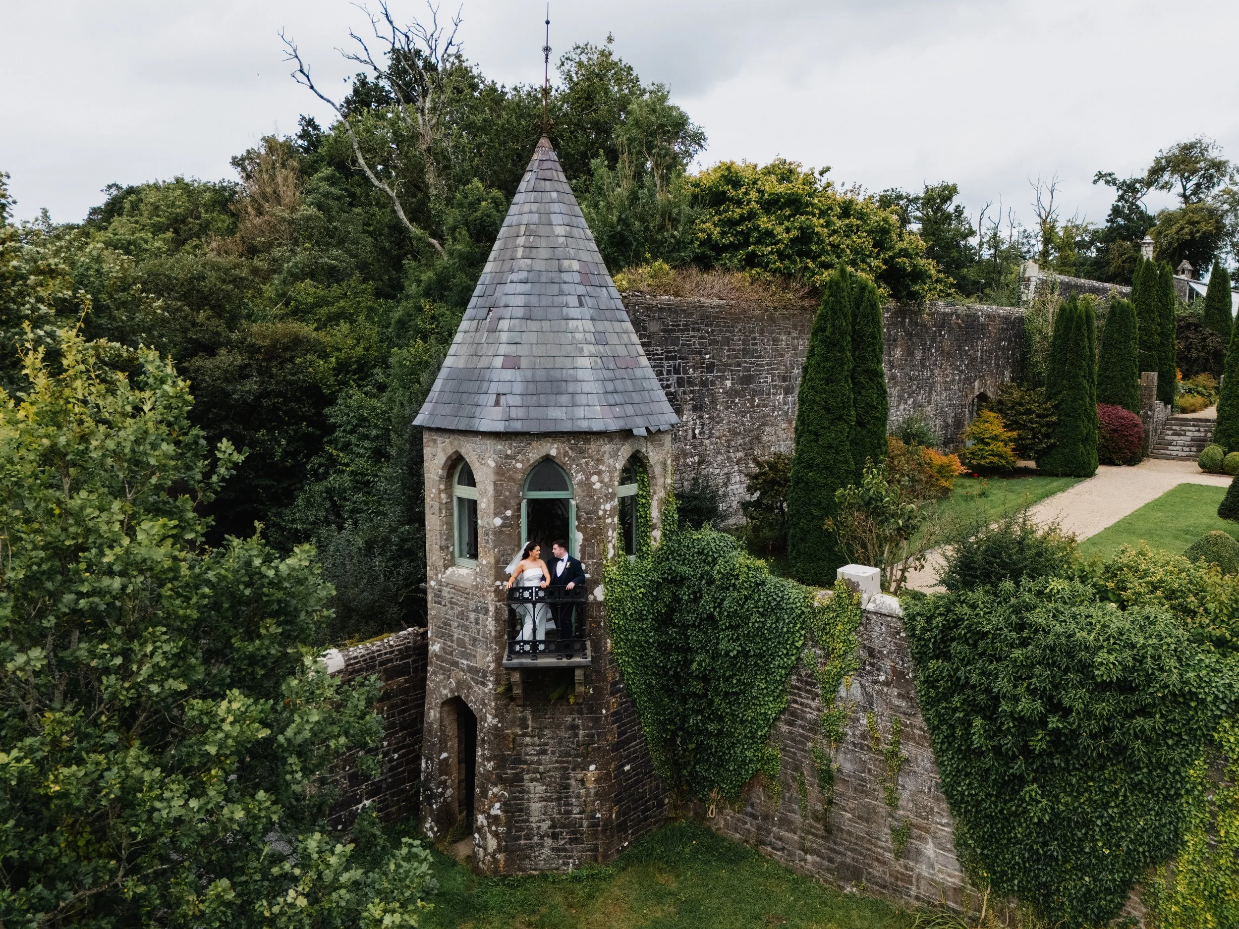 A small stone tower with a conical slate roof and arched windows, situated in lush green surroundings with trees and a stone wall.