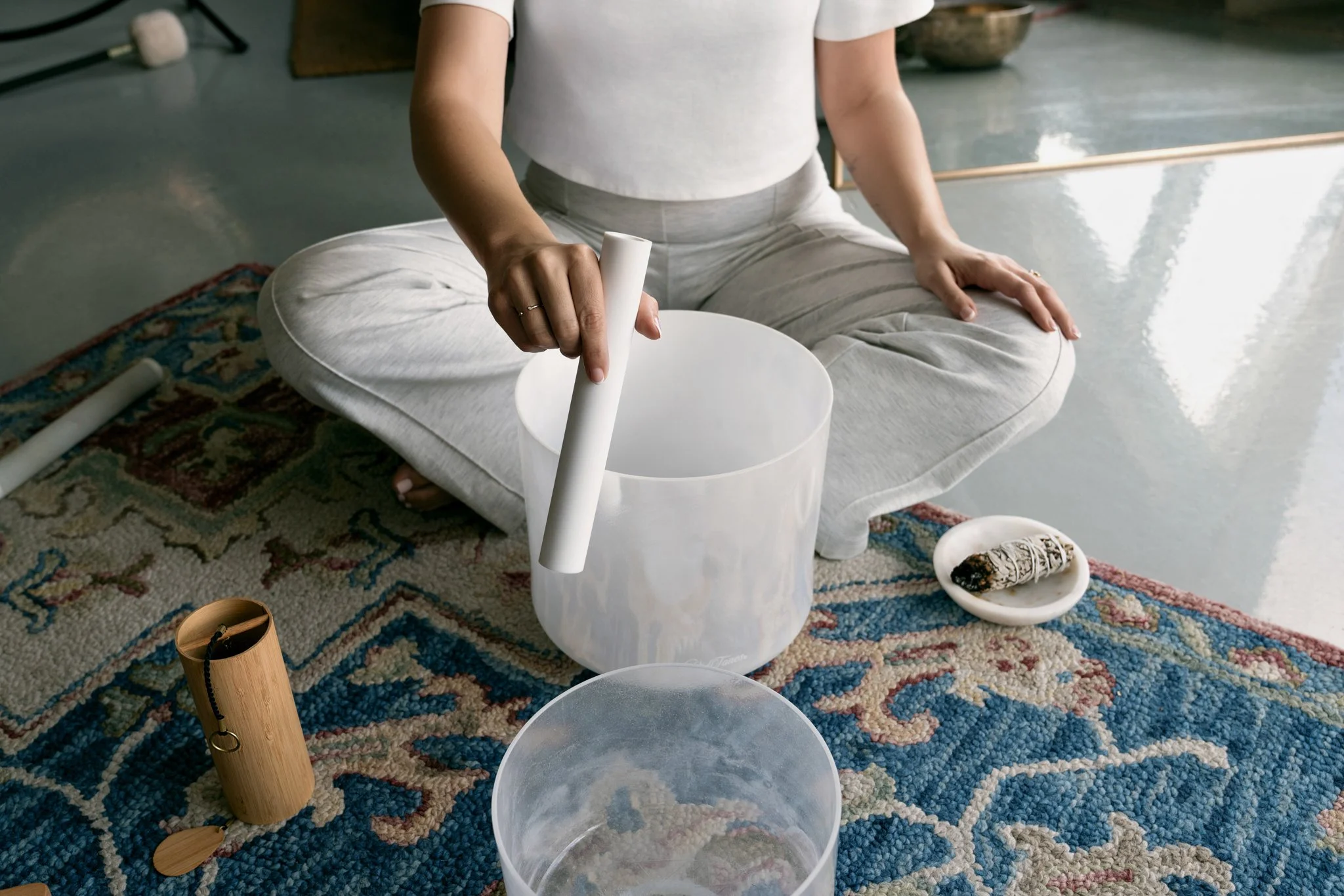 Person sitting cross-legged on a patterned rug, holding a mallet over a clear crystal singing bowl, with a plate of sage and other sound bowls nearby in a calming, minimalist space.
