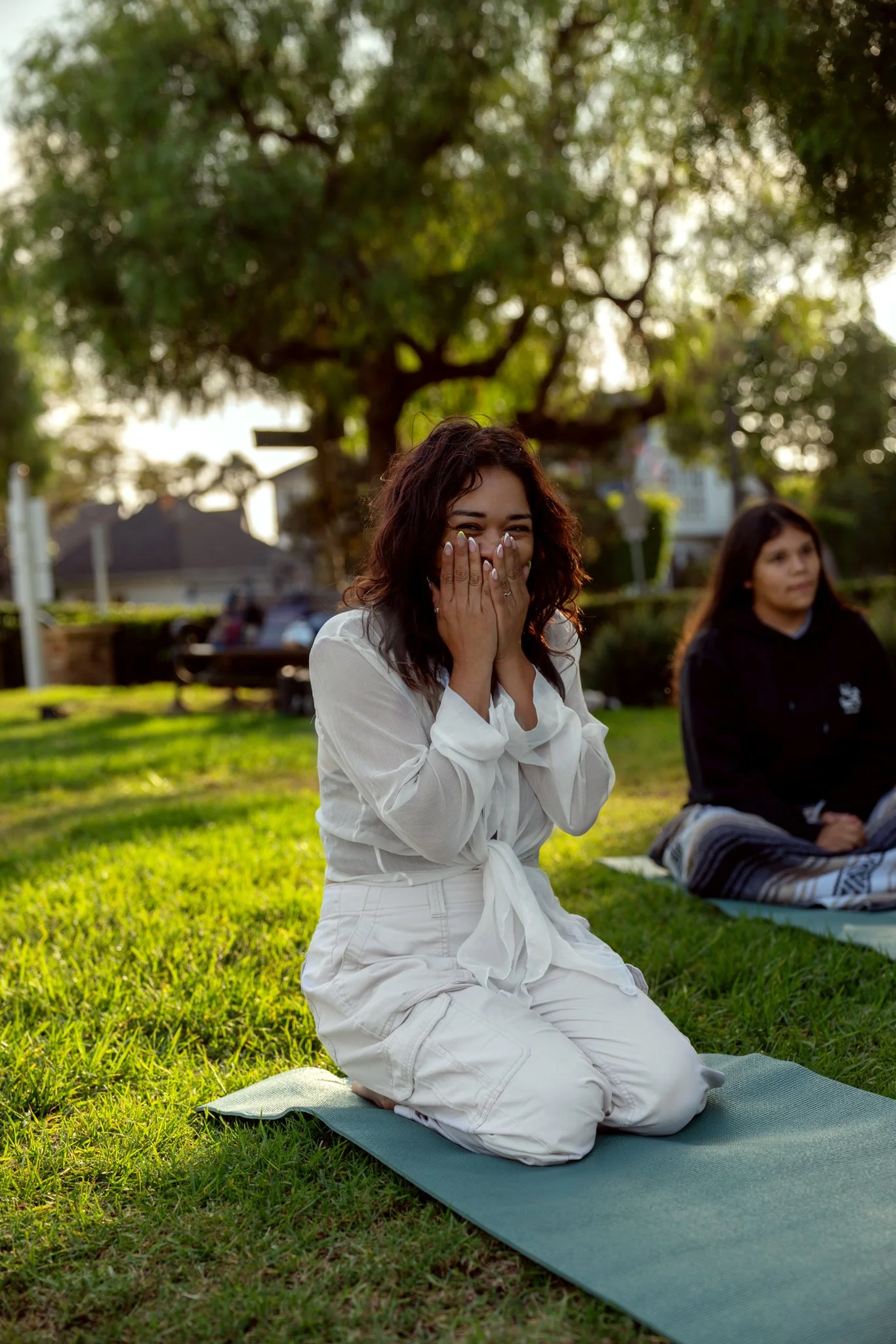 A woman with brown curly hair in white clothing sitting kneeling on a yoga mat outdoors, covering her mouth and nose with her hands, smiling, with another woman in a black hoodie sitting on a blanket nearby, under a large tree in a park during sunset.