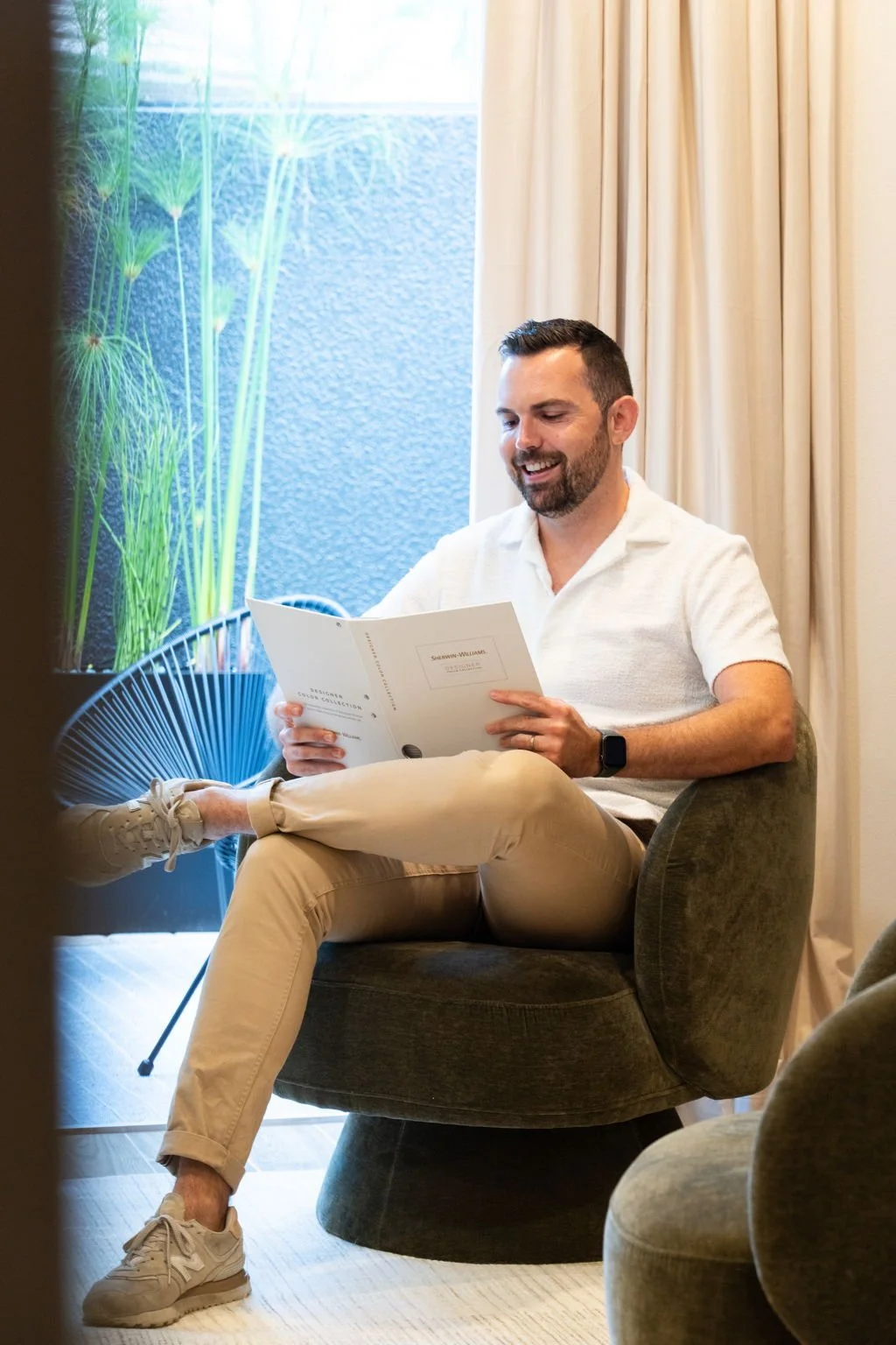 A man with dark hair and a beard is sitting in a comfortable armchair, smiling while reading a brochure or magazine, in a well-lit room with beige curtains and a window with some greenery outside.